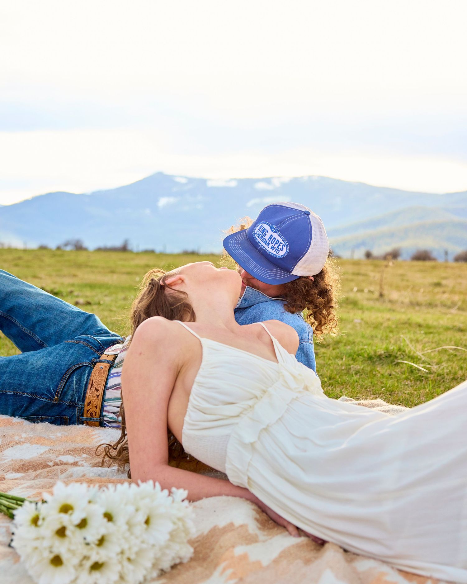 Romantic couple kissing on blanket in mountain field, bride in white dress with daisy bouquet nearby.
