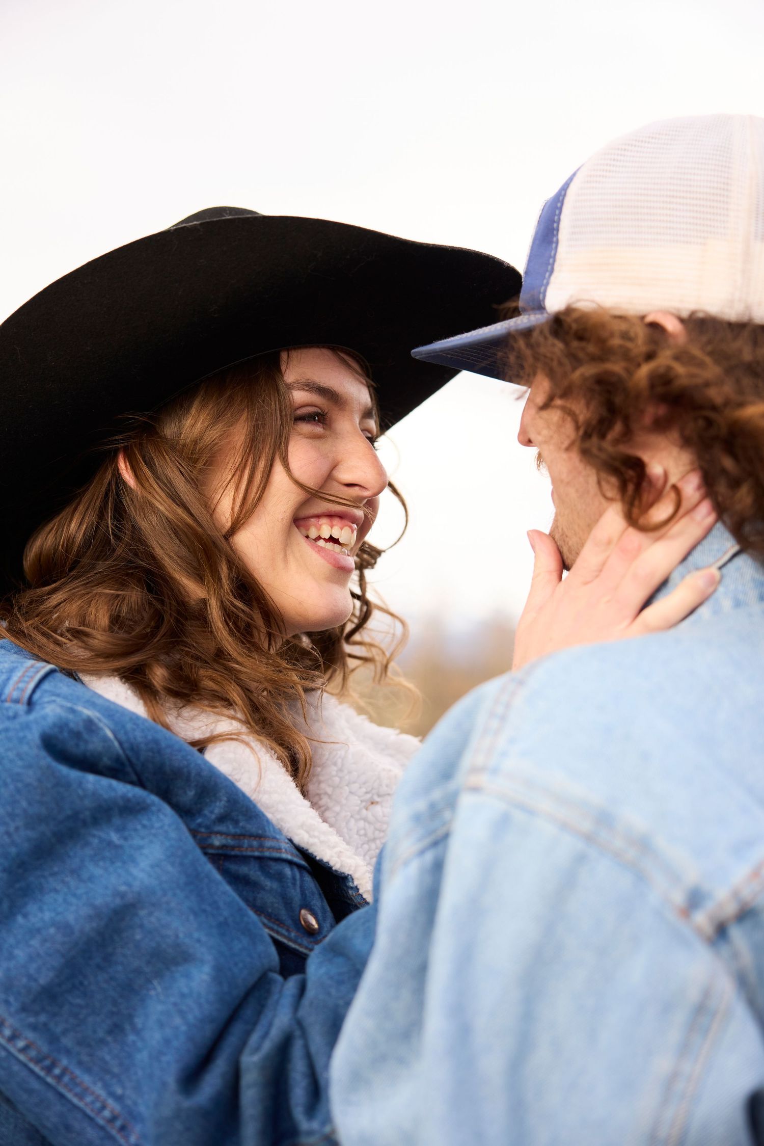 Smiling couple in denim jackets sharing a romantic moment, woman wearing black cowboy hat outdoors.
