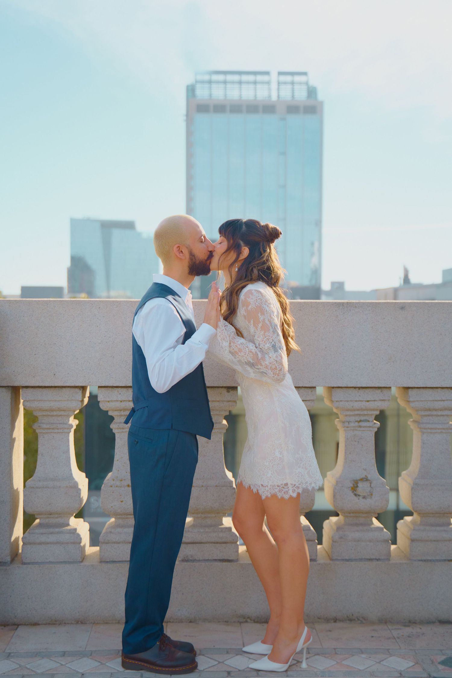 Newlyweds share a kiss on a rooftop terrace with city skyscrapers in the background.