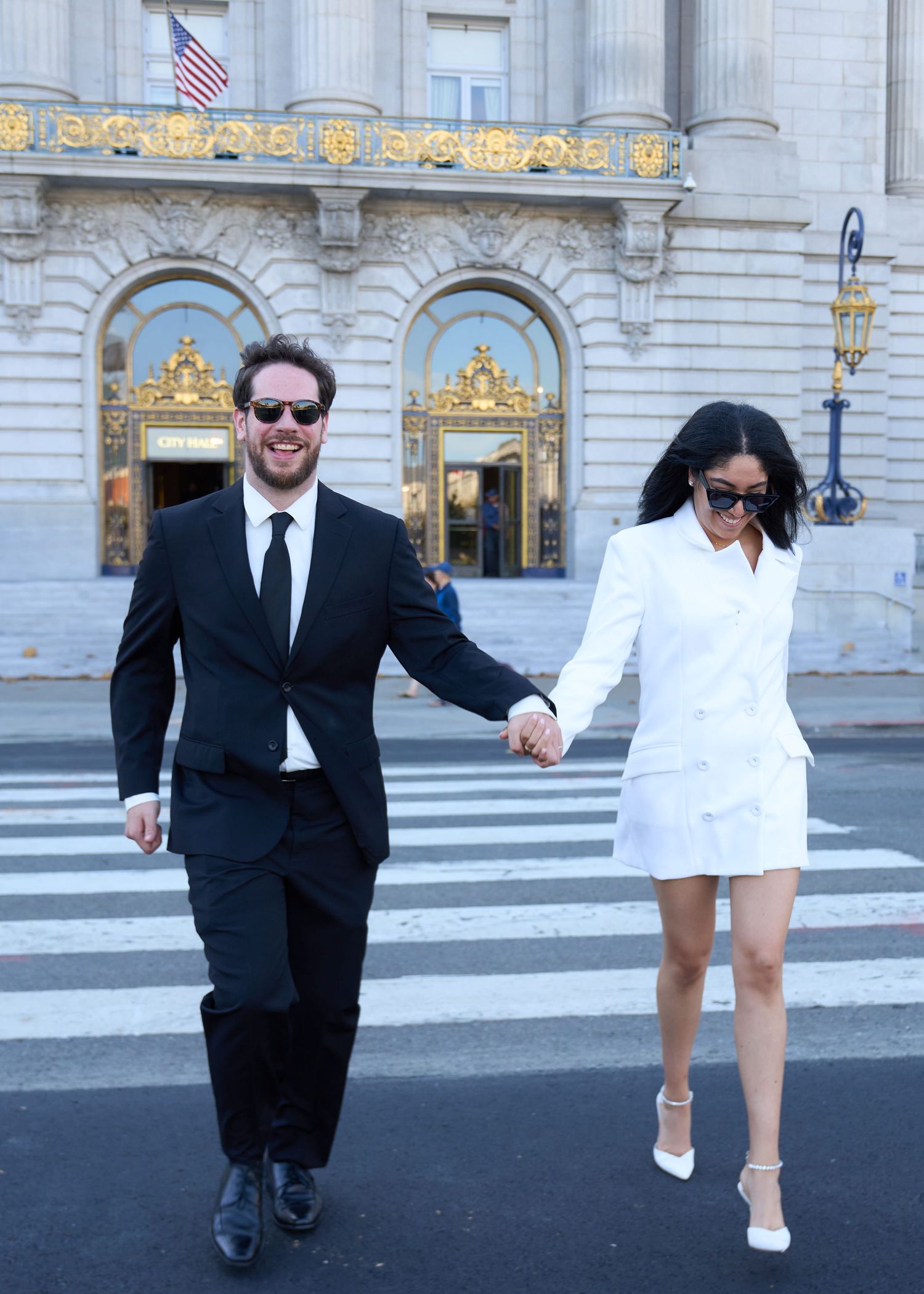 Smiling newlywed couple in wedding attire holding hands crossing street in front of San Francisco City Hall.