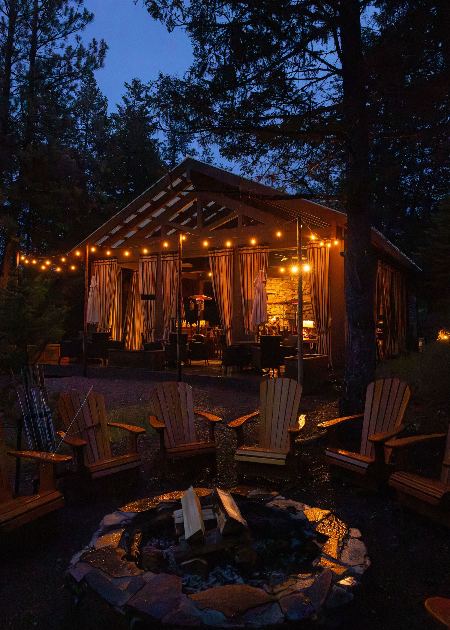 A cozy rustic cabin glows with warm string lights at dusk, surrounded by Adirondack chairs and a fire pit.