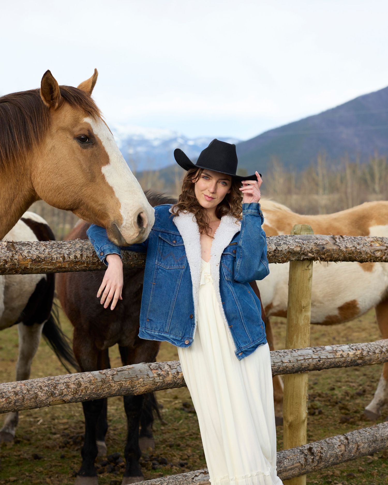 Woman in black cowboy hat and denim jacket posing beside a brown horse at a fence.