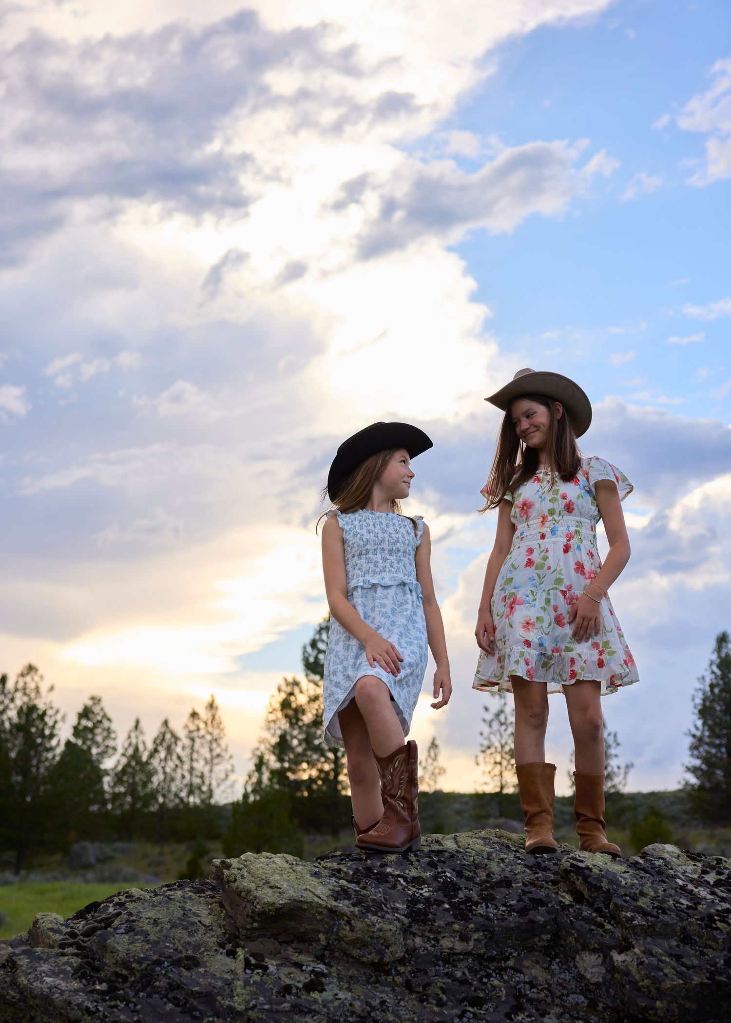 Two young girls wearing cowboy hats and dresses pose on a rock during a golden sunset.