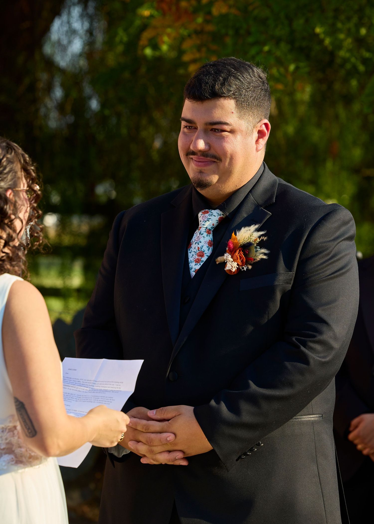 Groom in black suit with floral tie and boutonniere smiling during outdoor wedding ceremony while holding bride's hands.