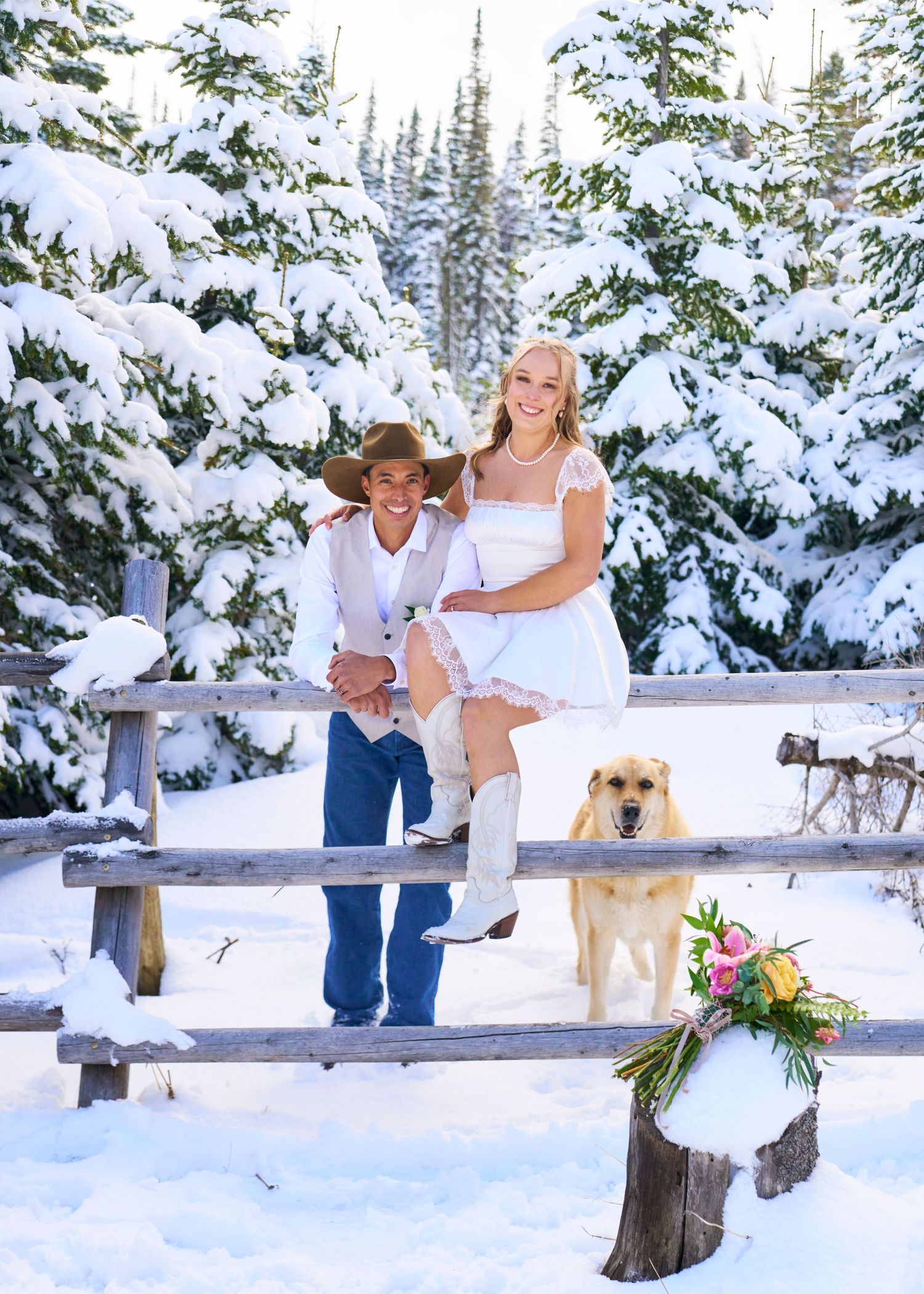 Couple in winter wedding attire pose on rustic fence with golden retriever amid snow-covered pine trees.