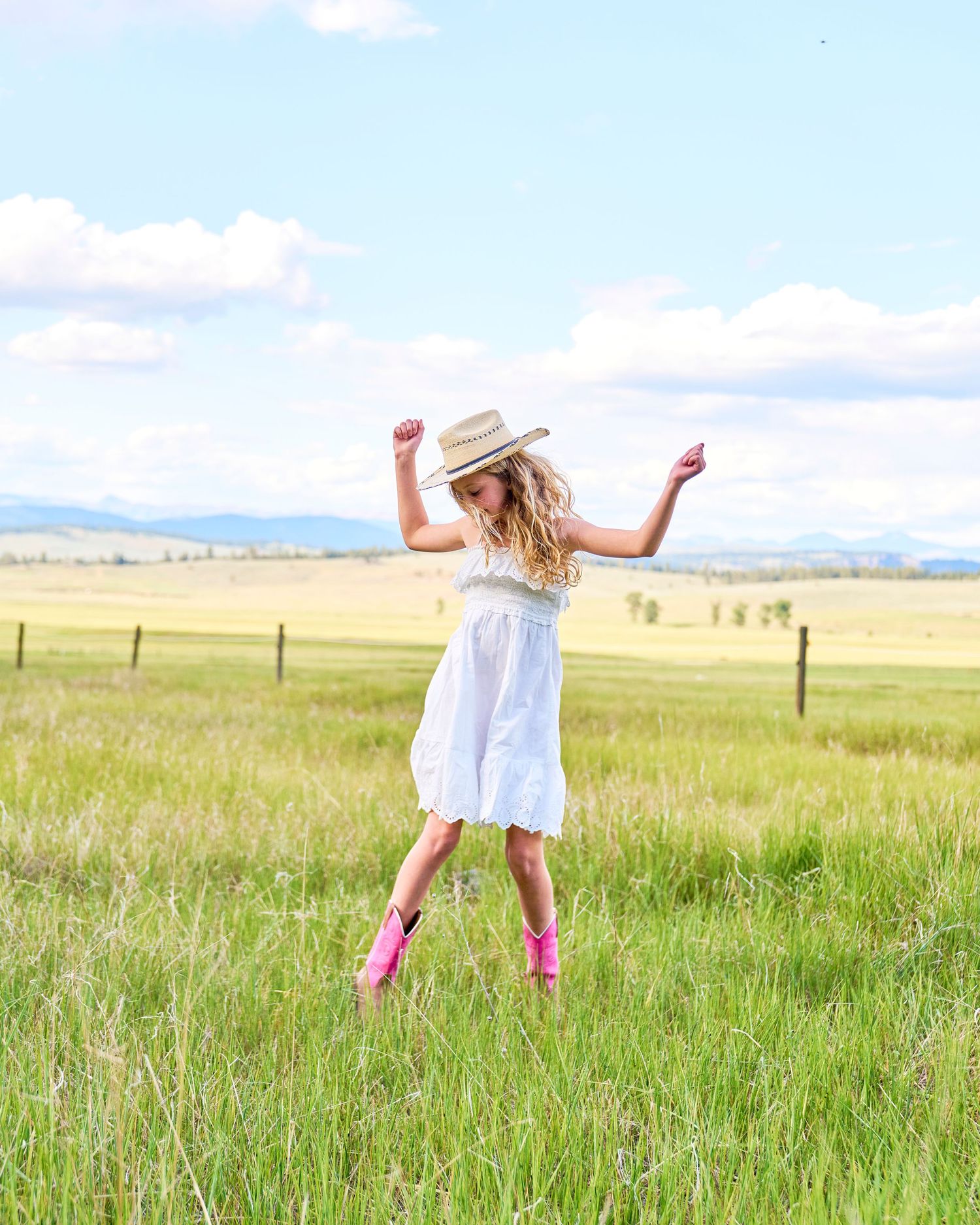 Young girl in white dress and straw hat dancing joyfully in a sunny green meadow with pink boots.