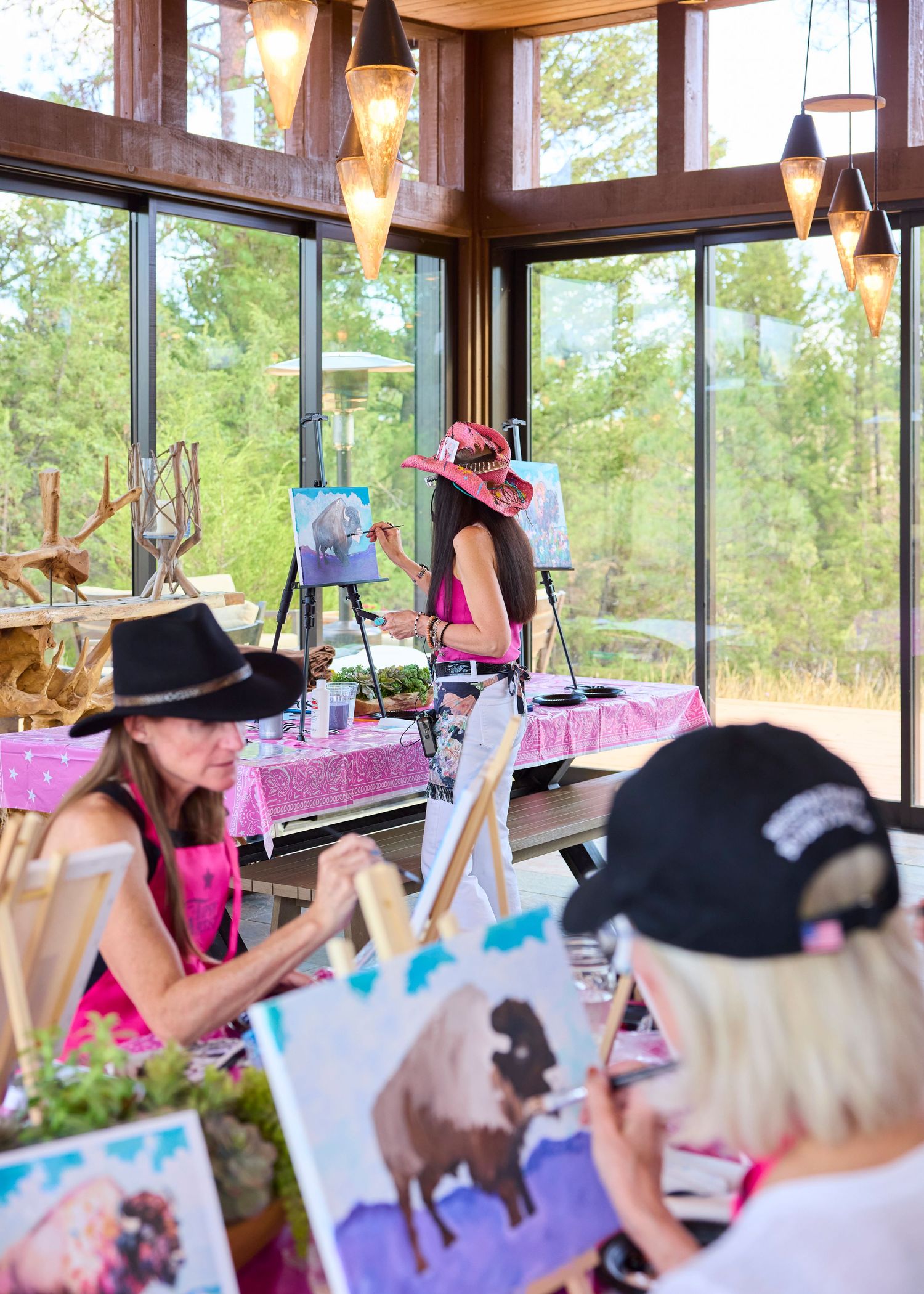 Women in pink aprons painting on easels in a rustic art studio with large windows overlooking nature.