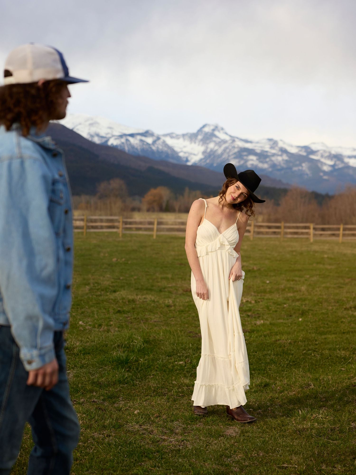 Woman in white dress and cowboy hat posing in a field with snow-capped mountains behind her.