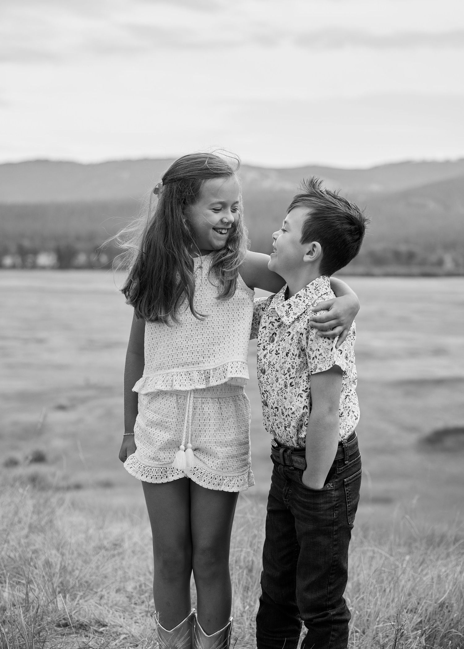 Black and white photo of two children smiling at each other in an open field with mountains behind.