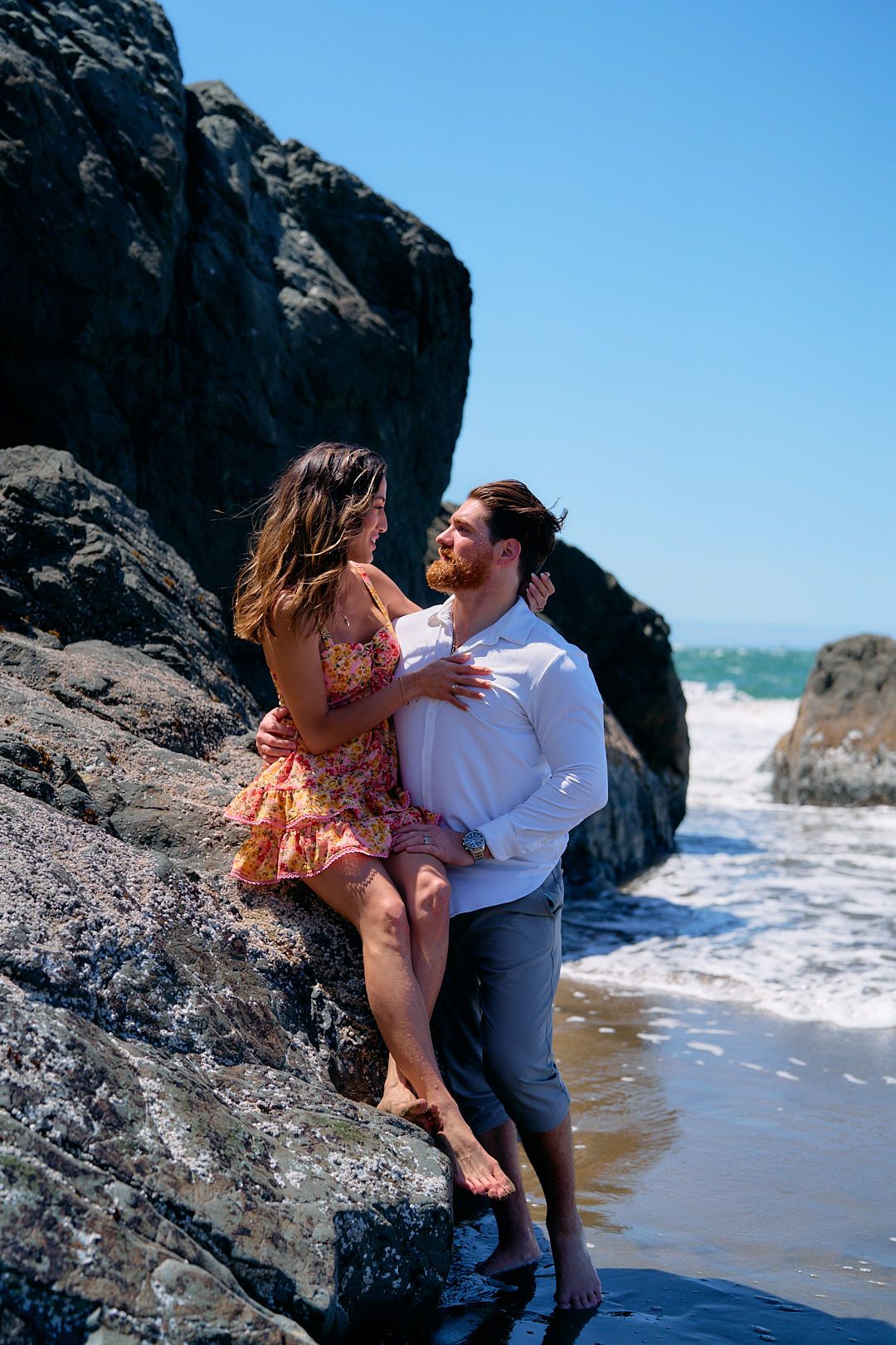 A couple shares a romantic moment on a rocky beach, woman sitting on a boulder, man in white shirt smiling.