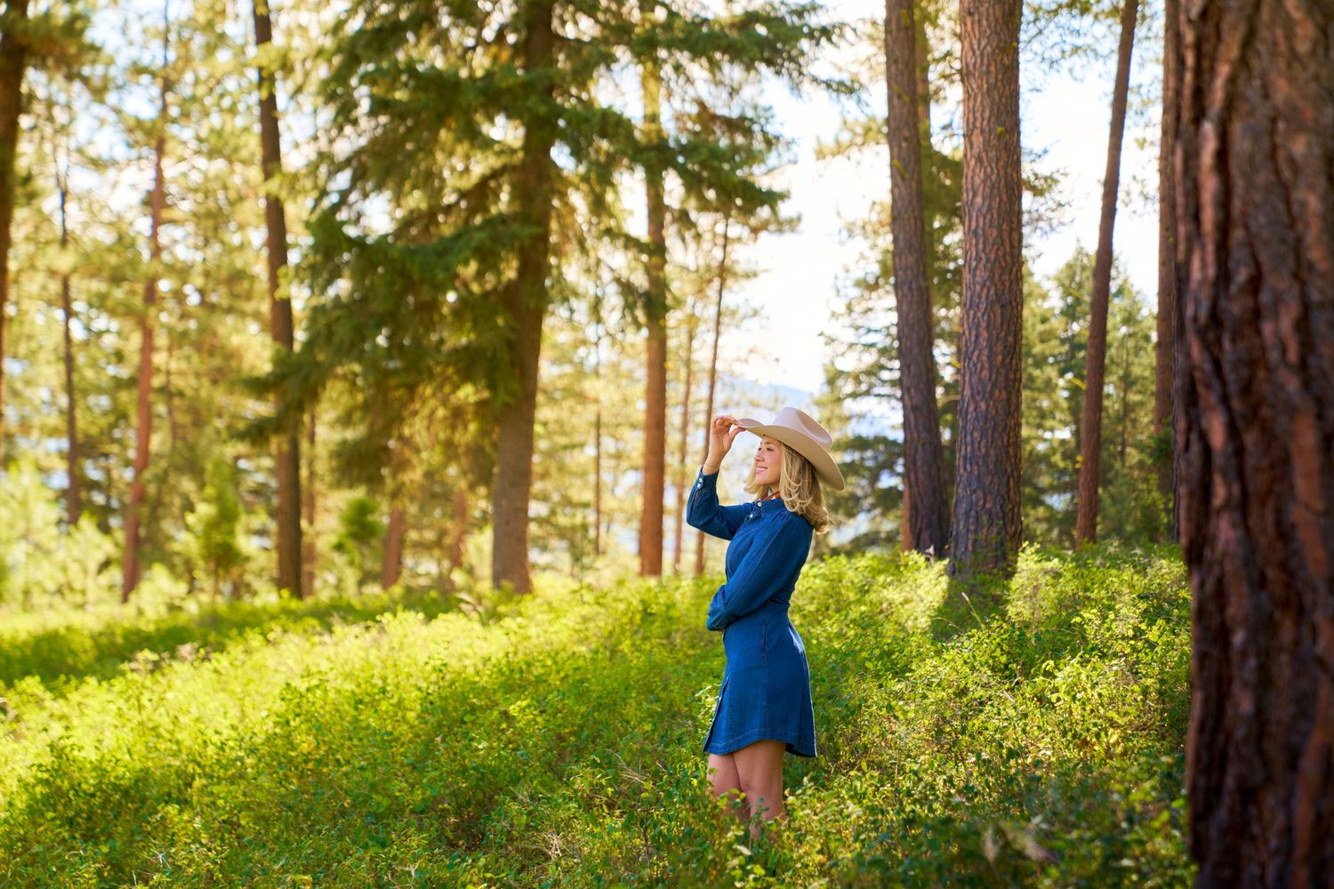 Blonde woman in a blue Pendleton dress and Stetson hat poses among tall pine trees in a sunlit forest.