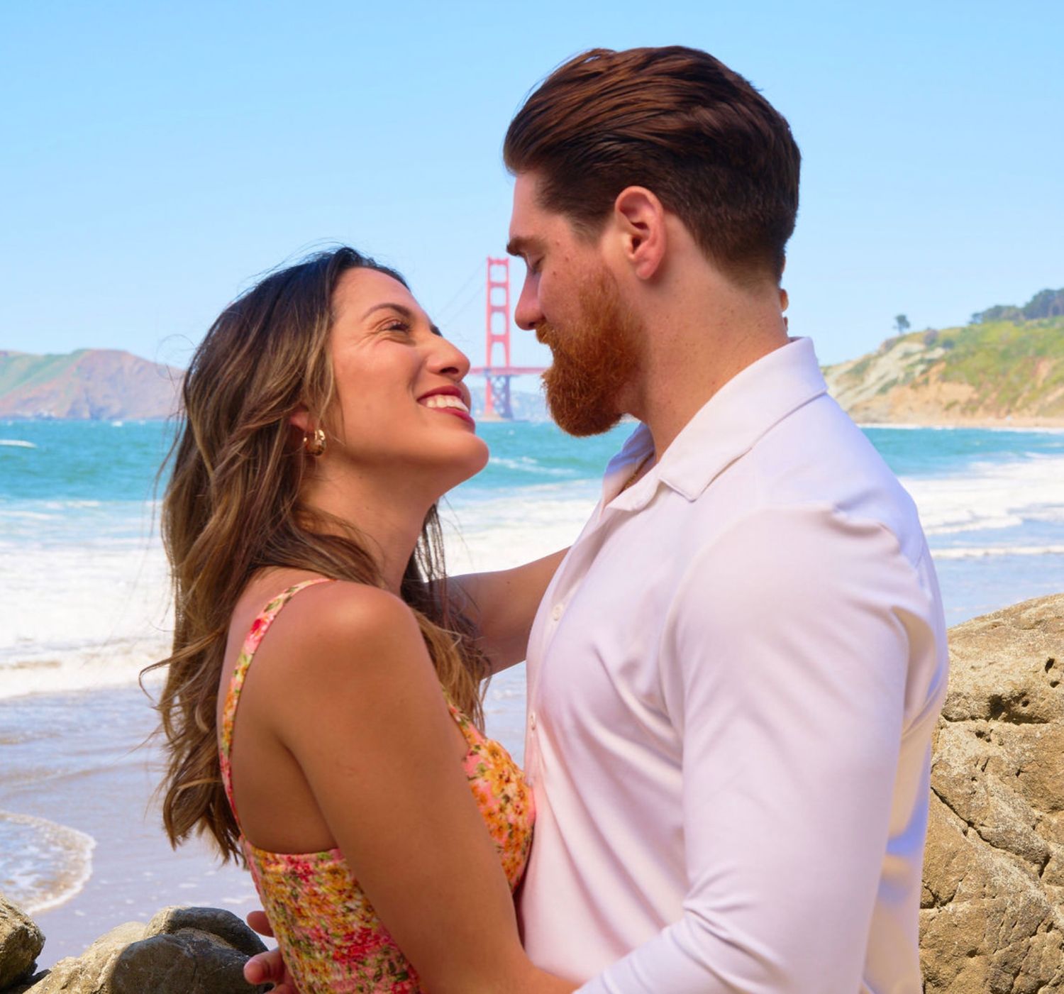 Smiling couple facing each other on Baker Beach with Golden Gate Bridge in background.