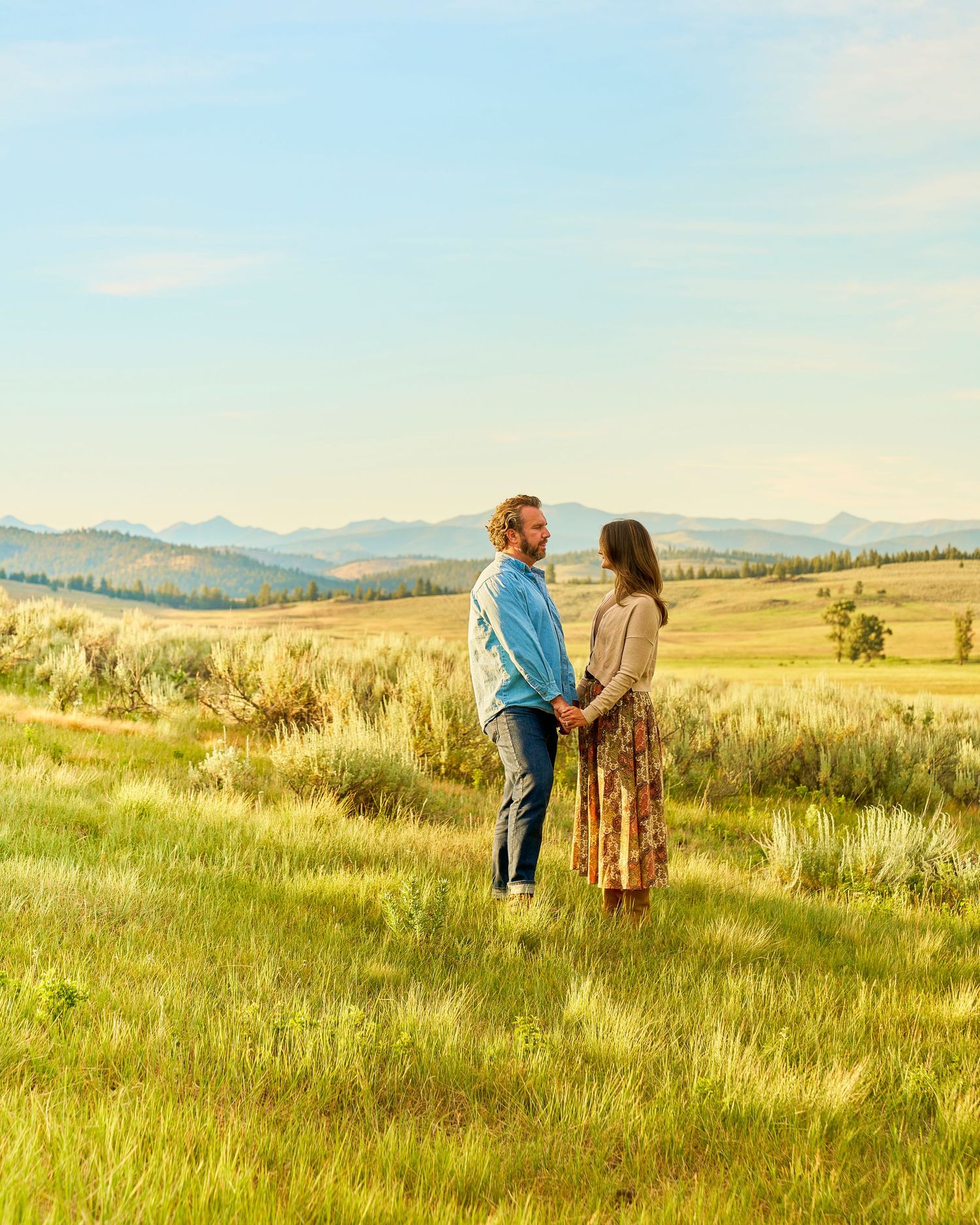 Couple faces each other holding hands in lush green field with mountain landscape behind them.