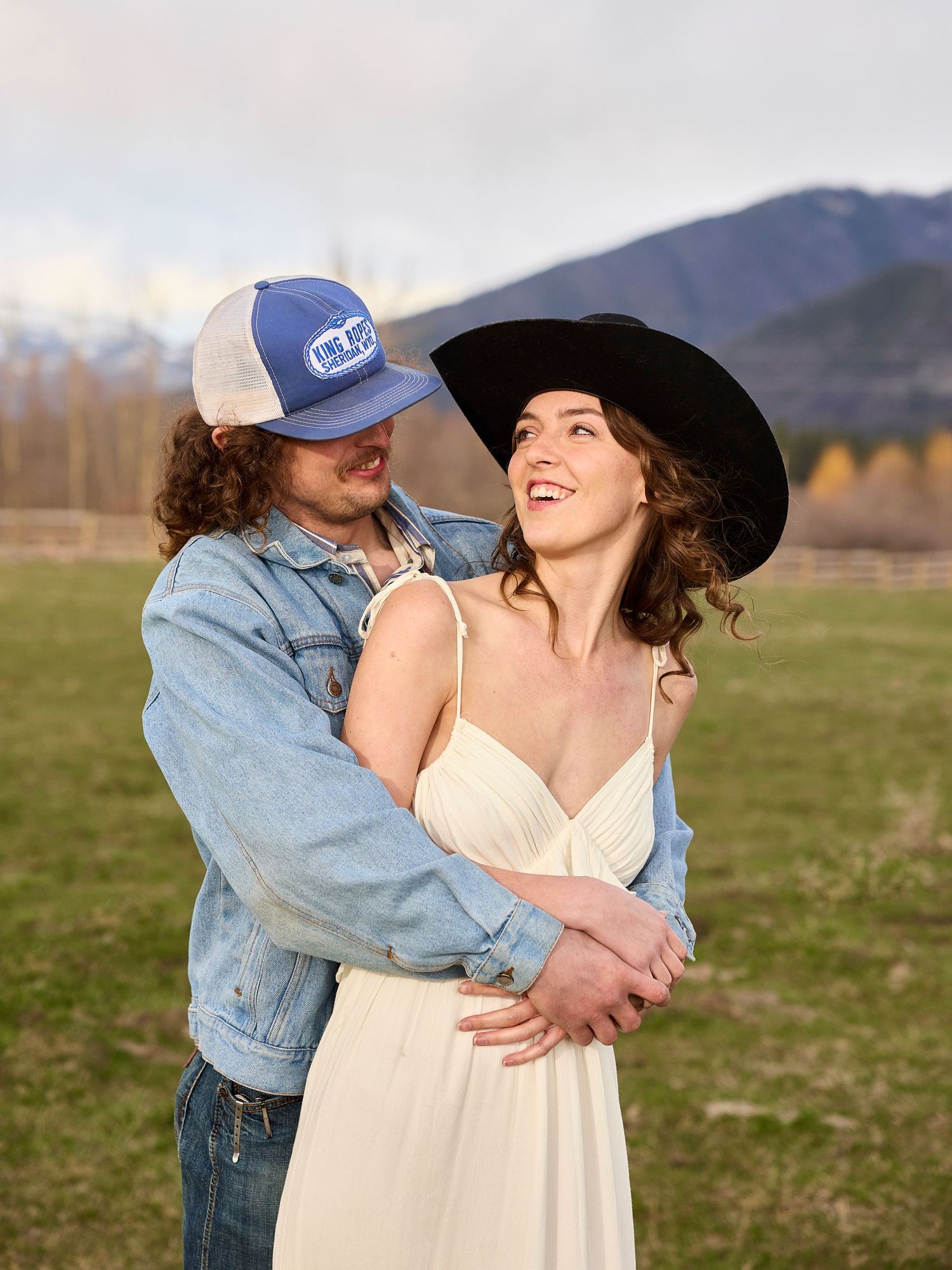 Couple embraces in field, woman in white dress and black hat, mountains in background.