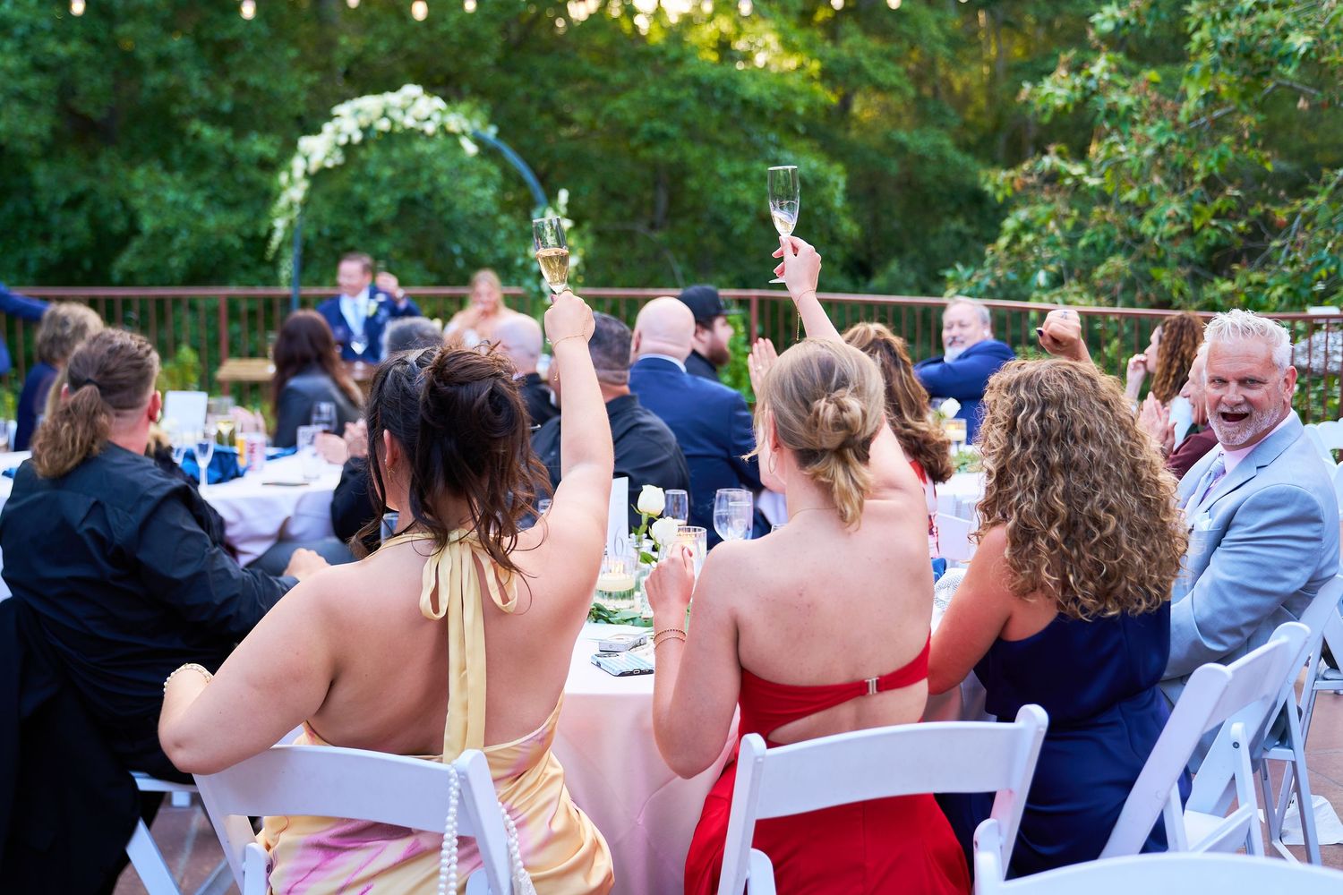 Wedding guests raising champagne glasses in a toast at an outdoor garden reception with string lights.