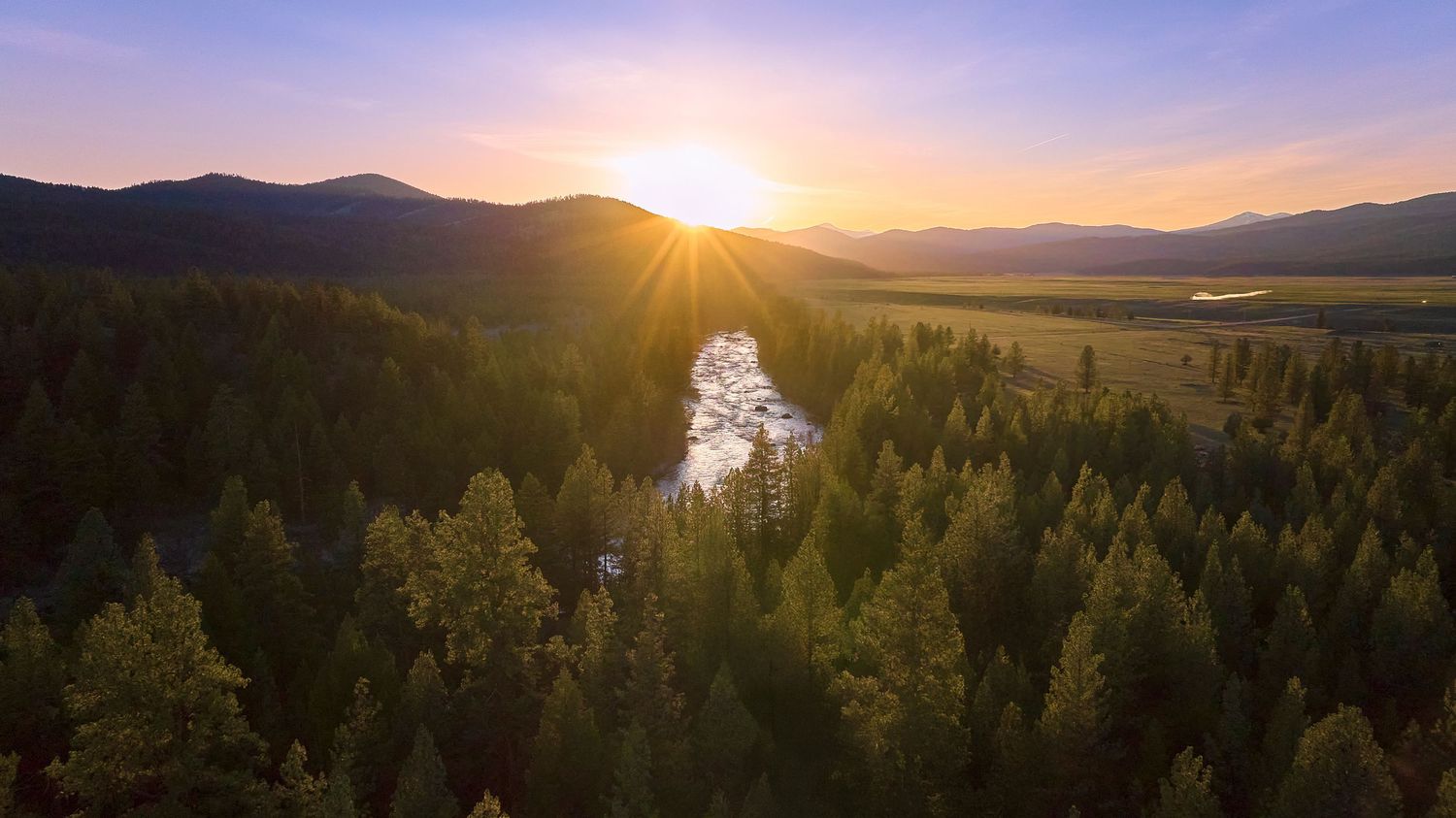 Aerial panoramic sunset over a forested river valley with warm sunburst rays and layered mountain silhouettes.