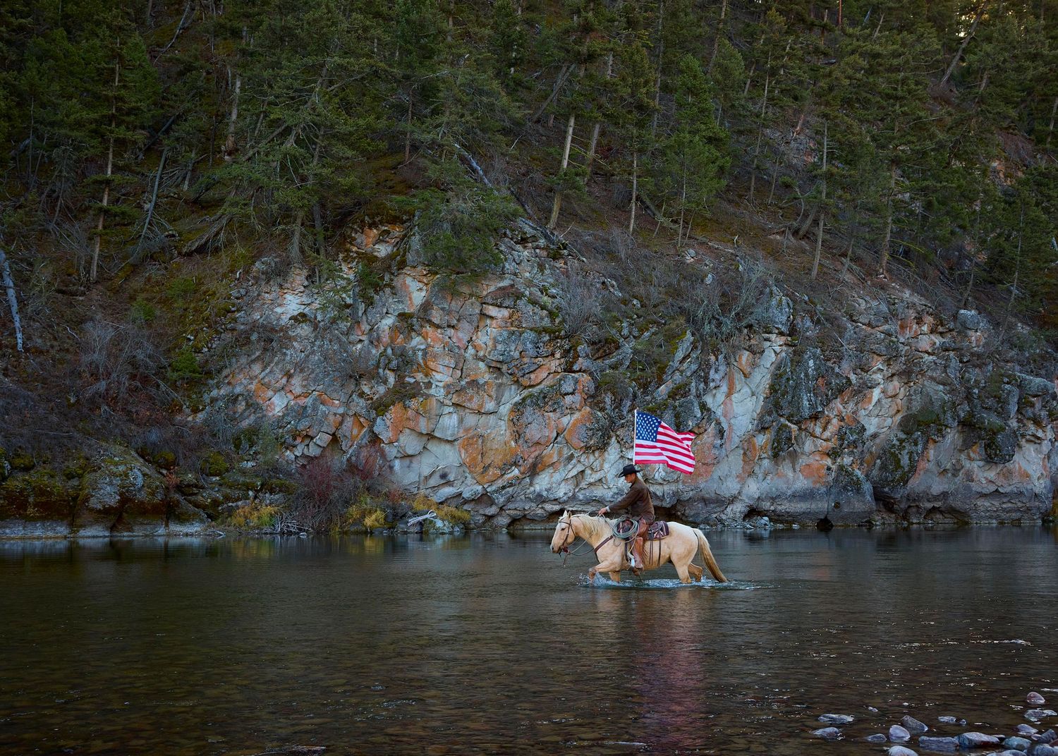 Rider on horseback carrying an American flag crosses a river surrounded by rocky forested terrain.
