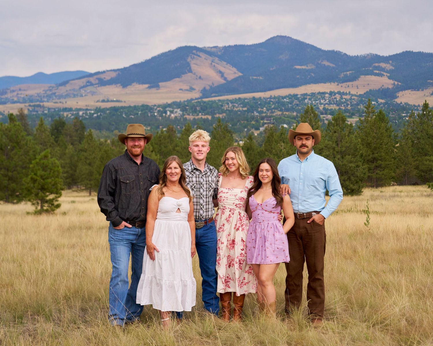 Family of six posing in open field with stunning Montana mountain range backdrop on cloudy day.