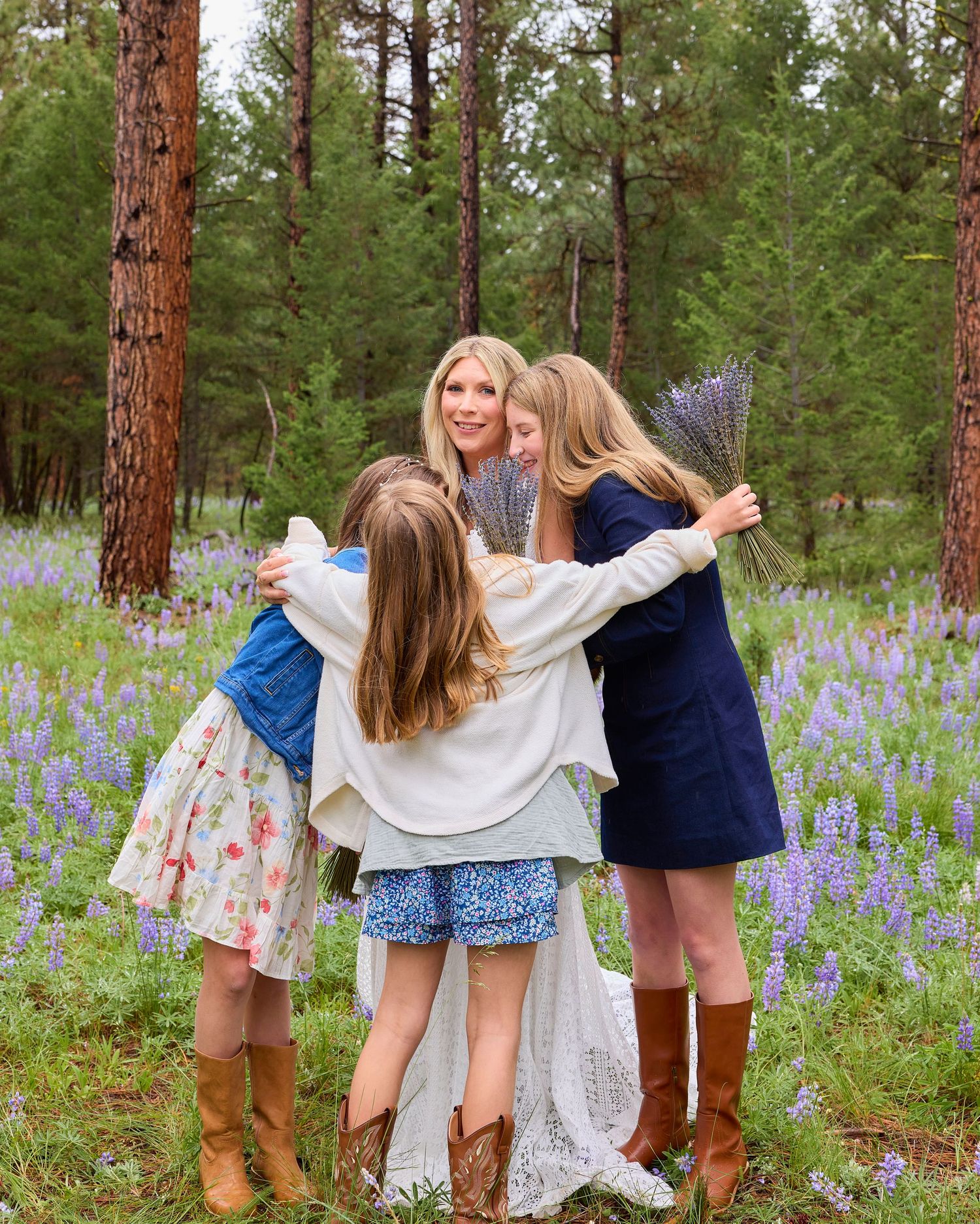 Bride in white lace gown embraces three girls in a purple lupine-filled pine forest during outdoor wedding.