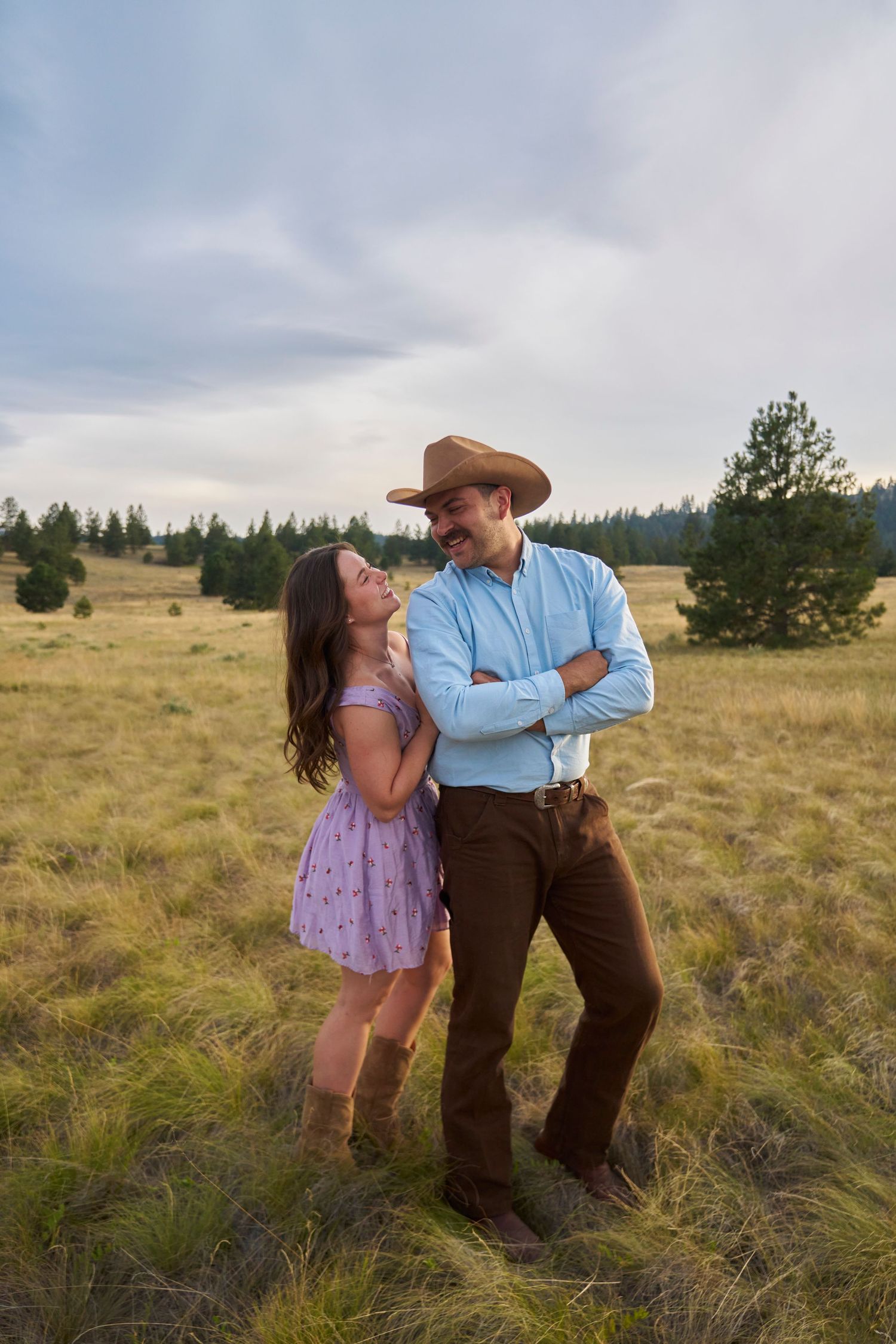 A couple in western attire shares a sweet moment in a golden grass field, man in cowboy hat with arms crossed.