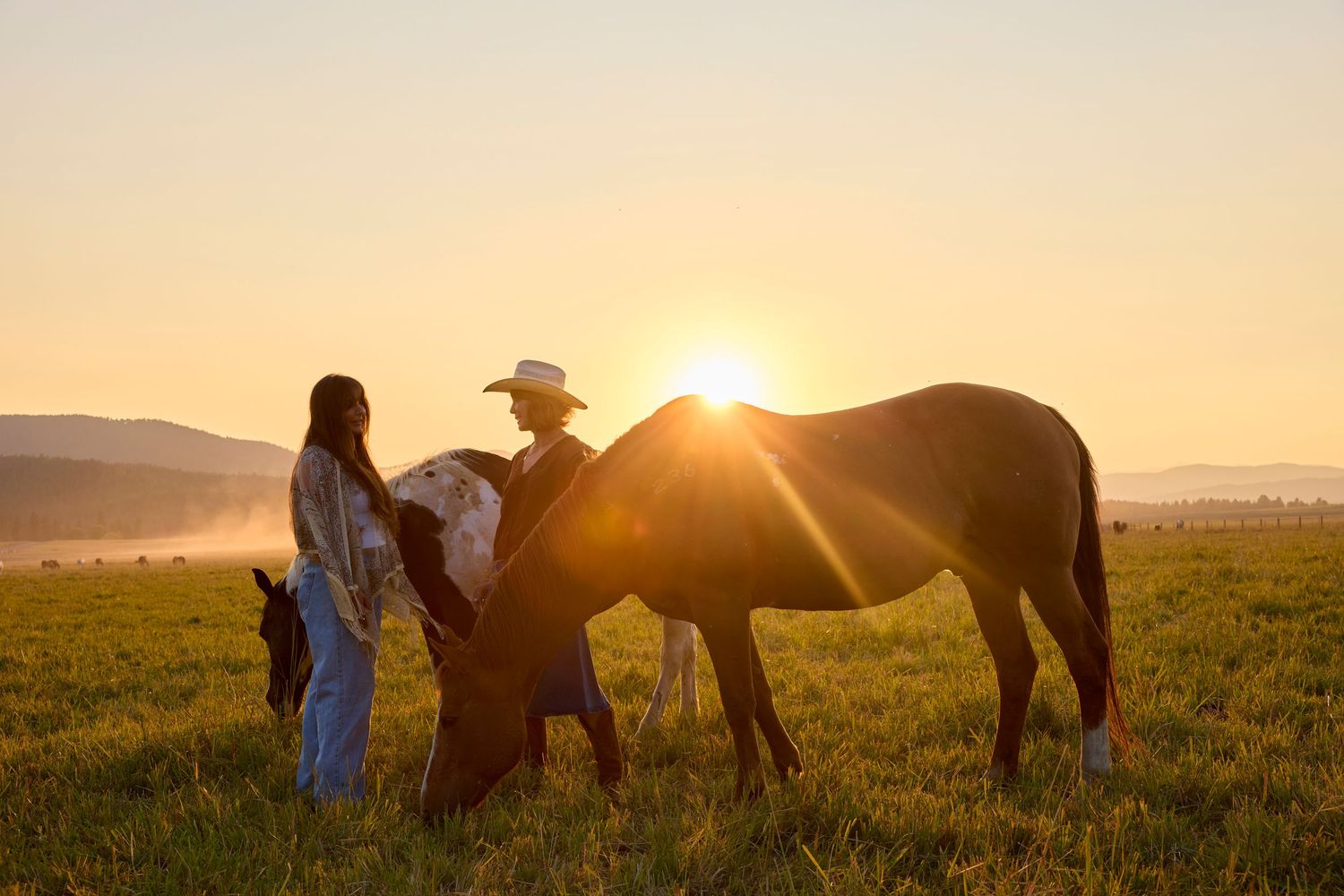 Couple standing with horses in open field during stunning golden sunset on a ranch.