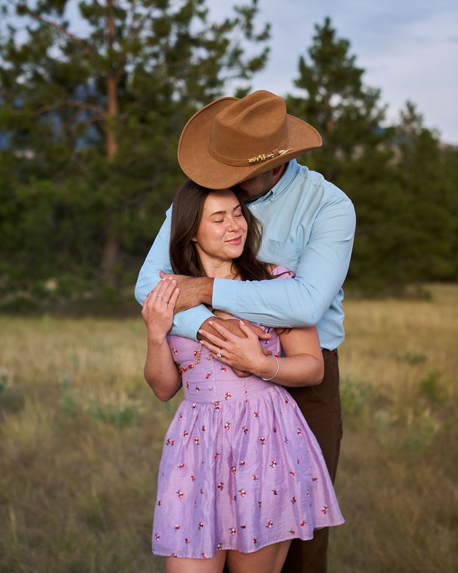 Couple embracing in a field, man in cowboy hat and blue shirt hugging woman in lavender floral dress.