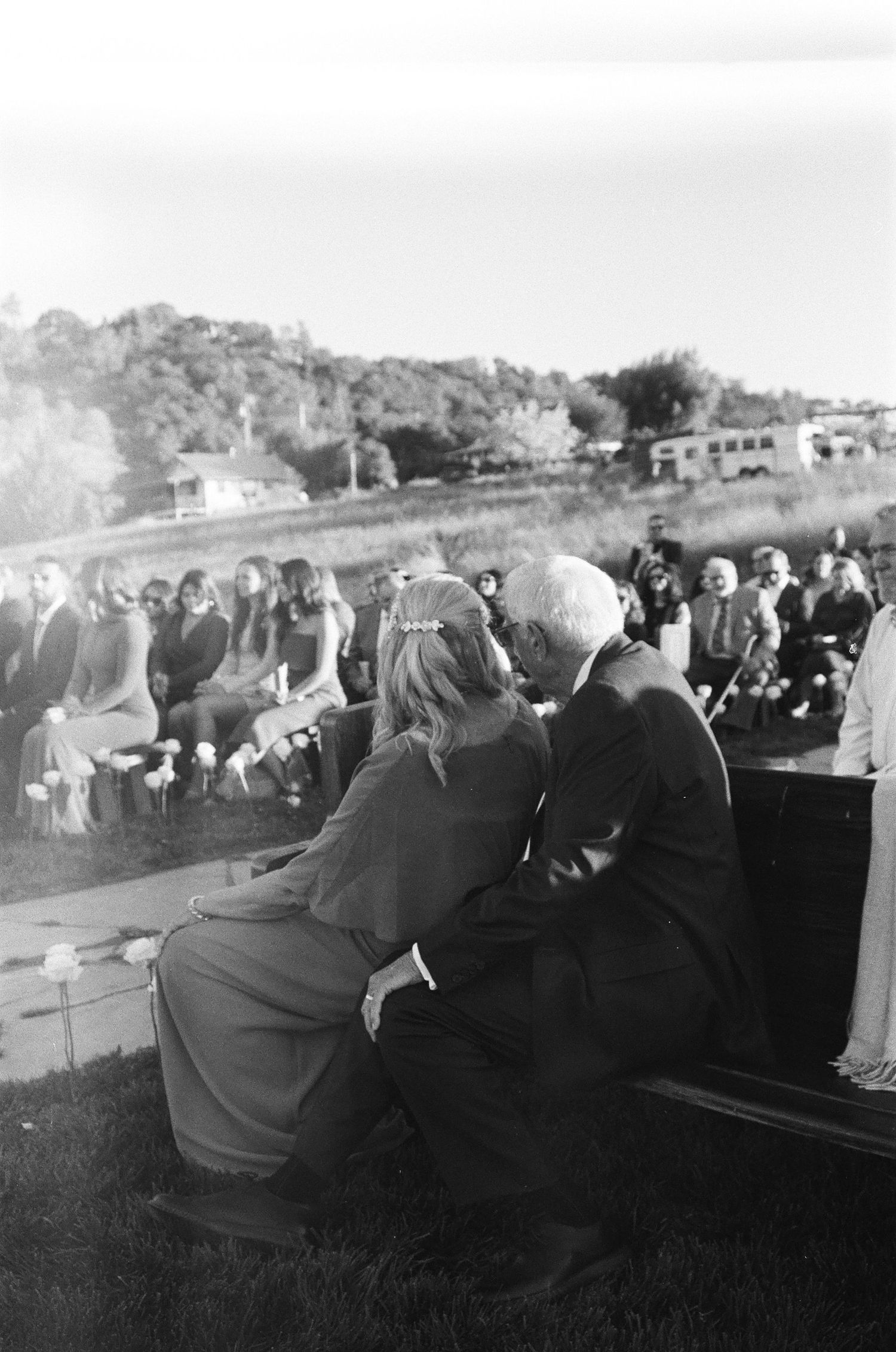 Couple sits closely together watching outdoor wedding ceremony with guests seated on hillside venue.