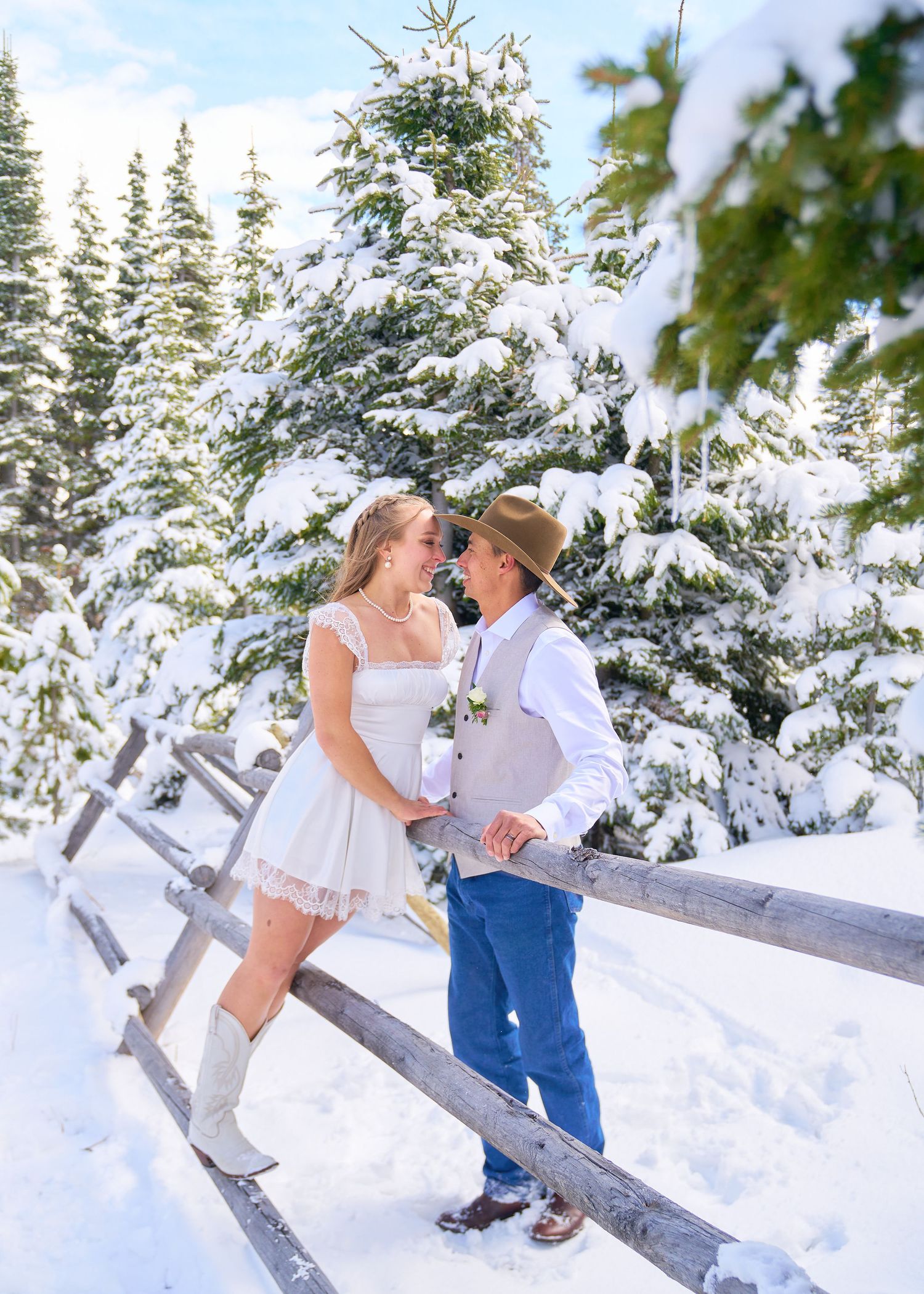 Bride in white mini dress and groom in cowboy hat touch foreheads by rustic fence in snowy forest.