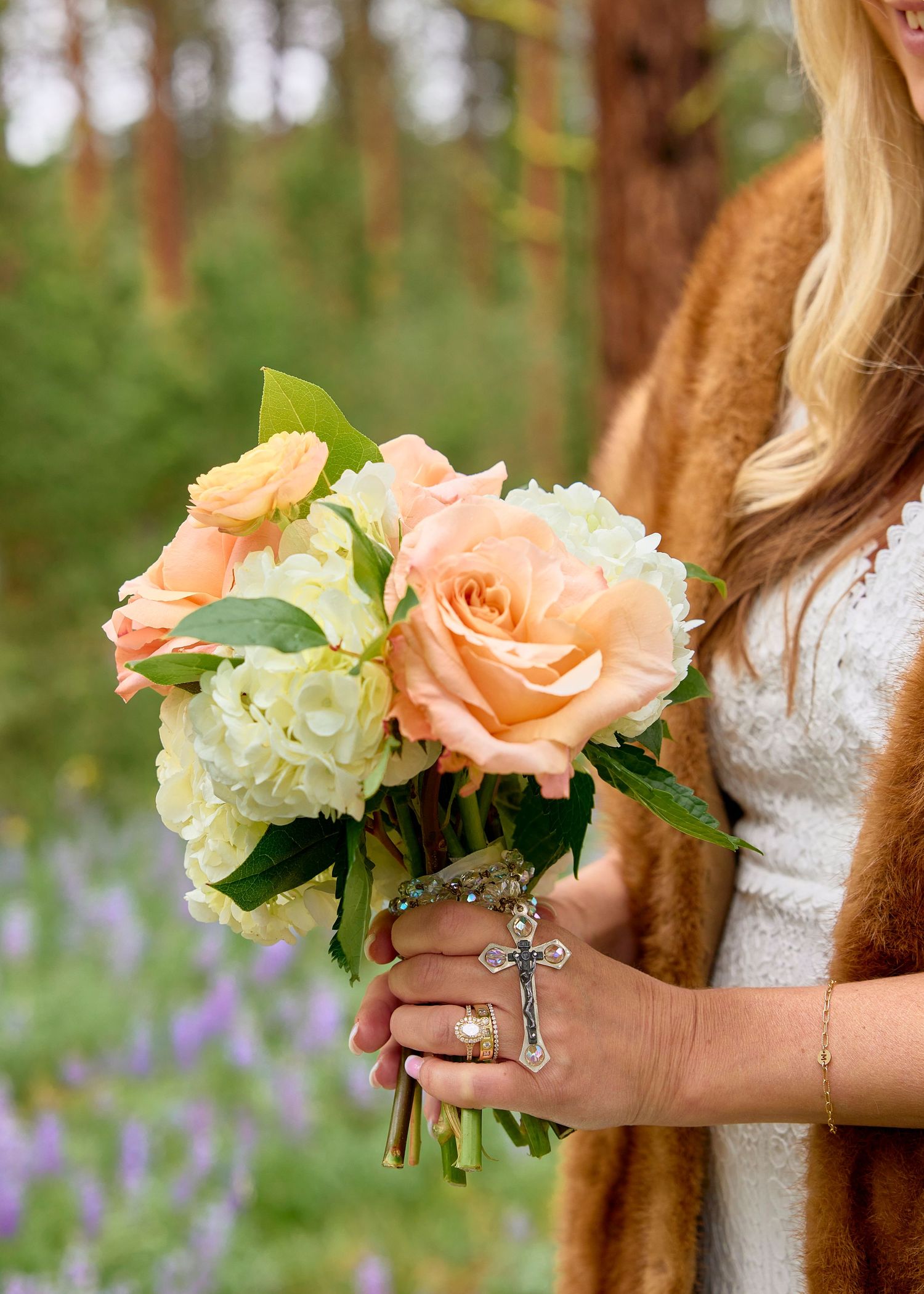 Bride holding peach rose and white hydrangea bouquet with rosary in a pine forest setting.