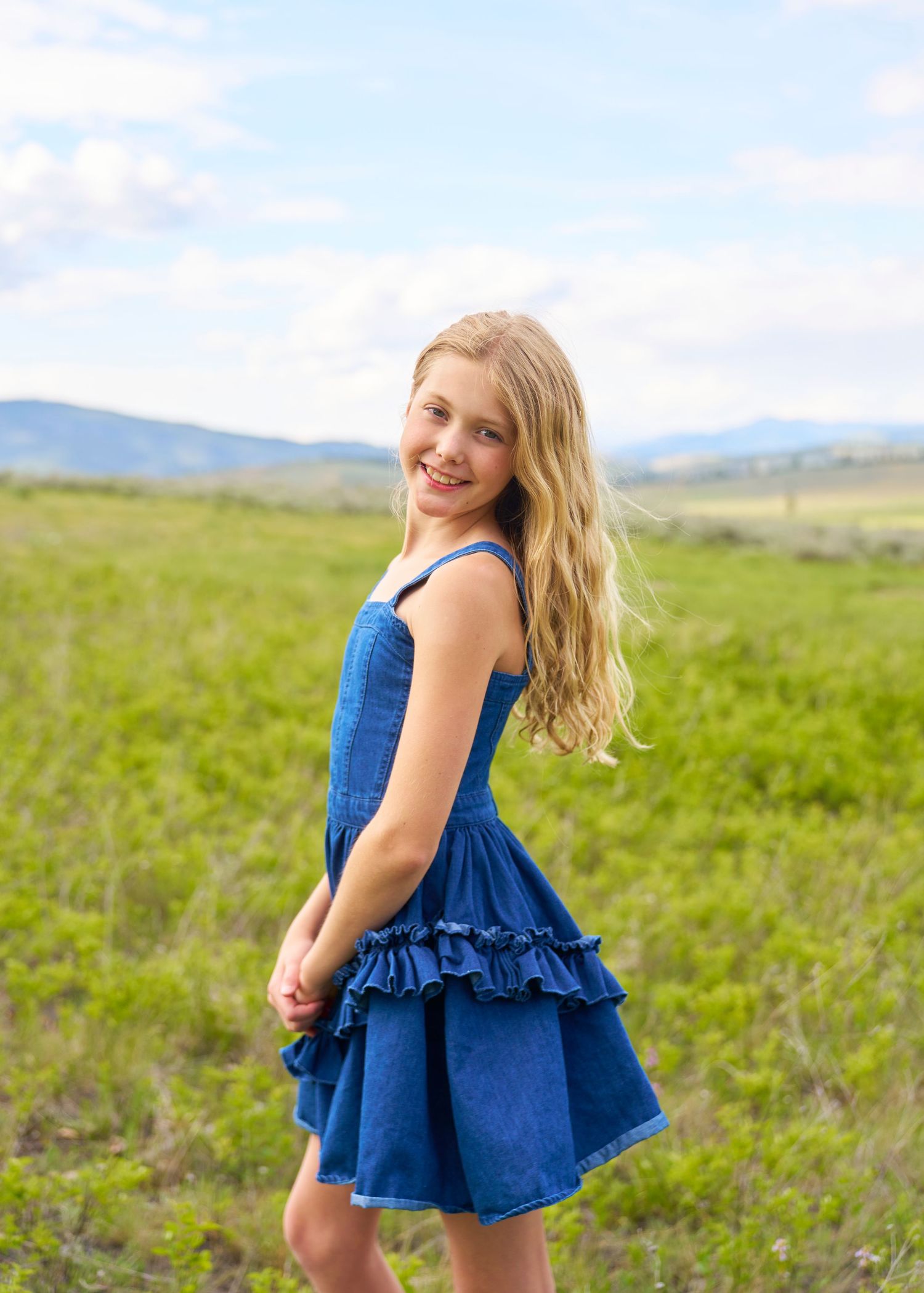 Smiling blonde girl in blue ruffled denim dress posing in green mountain meadow.