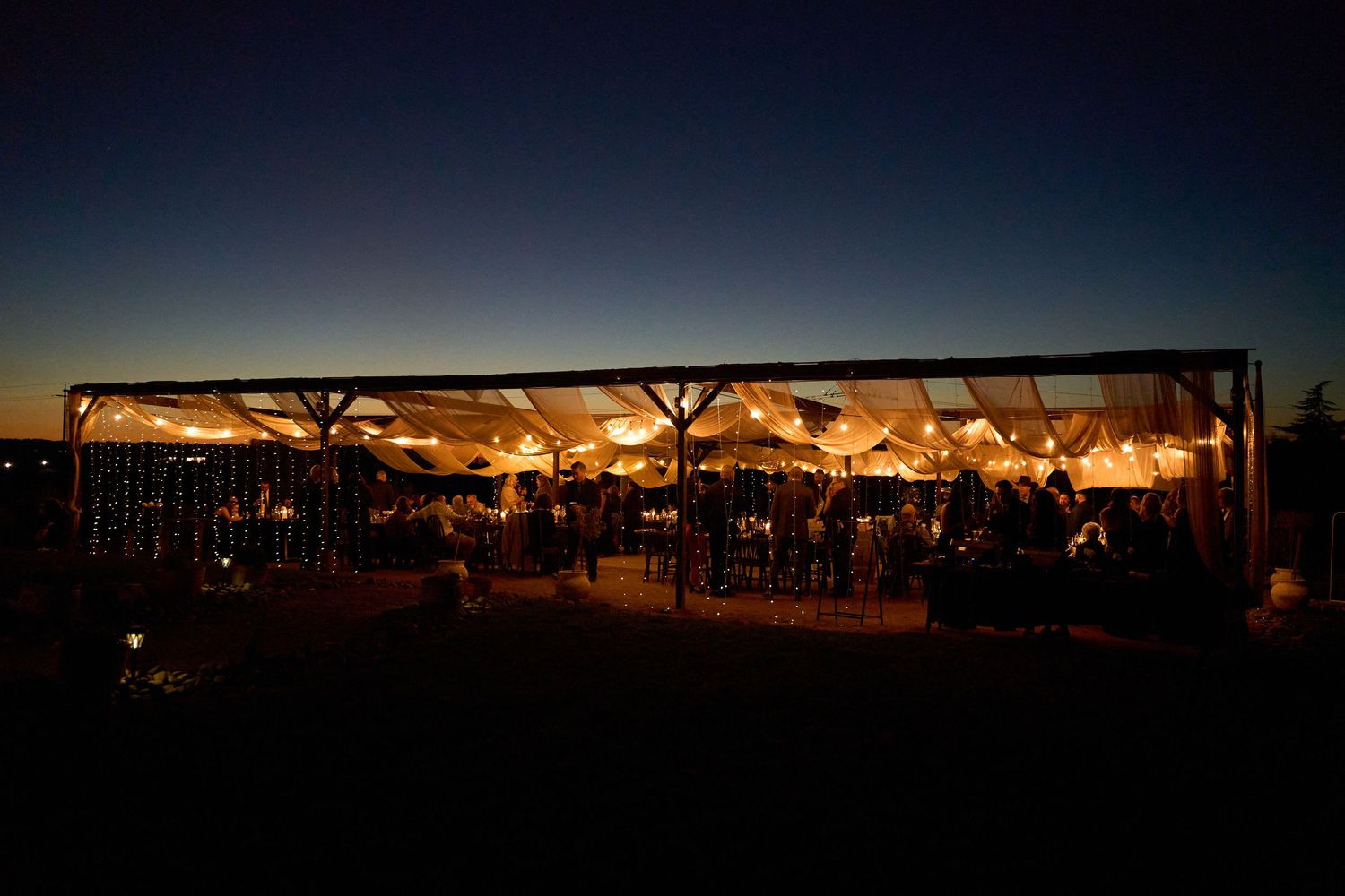 Stunning outdoor wedding reception tent glowing with warm lights against a deep blue twilight sky with guests dining inside.