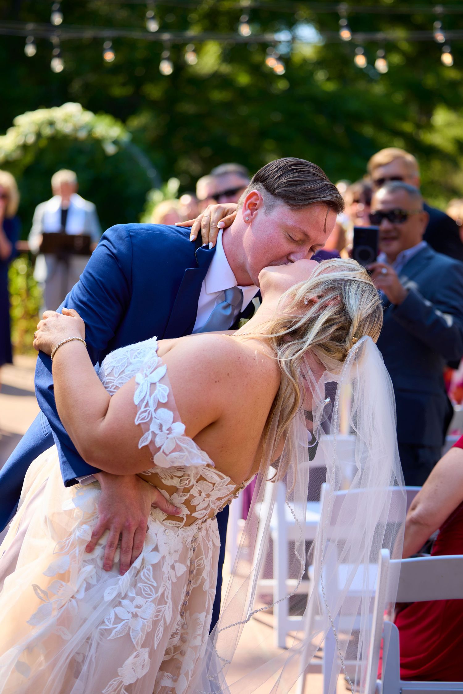Groom dips bride for first kiss at outdoor wedding ceremony with guests cheering in background.