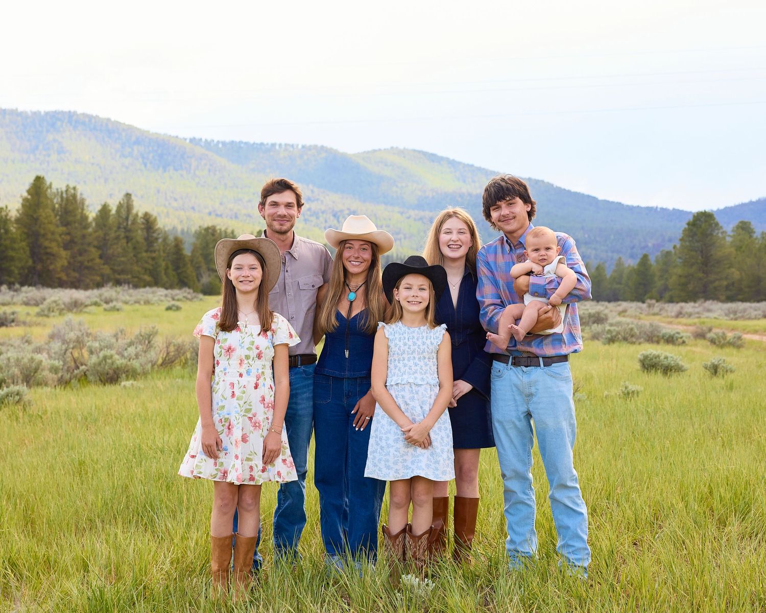 Family of seven smiling together in a lush green mountain meadow wearing casual western outfits.