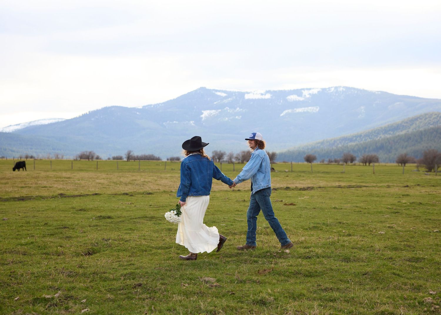 Couple walking hand in hand through a green field with snow-capped mountains and a cow in the background.