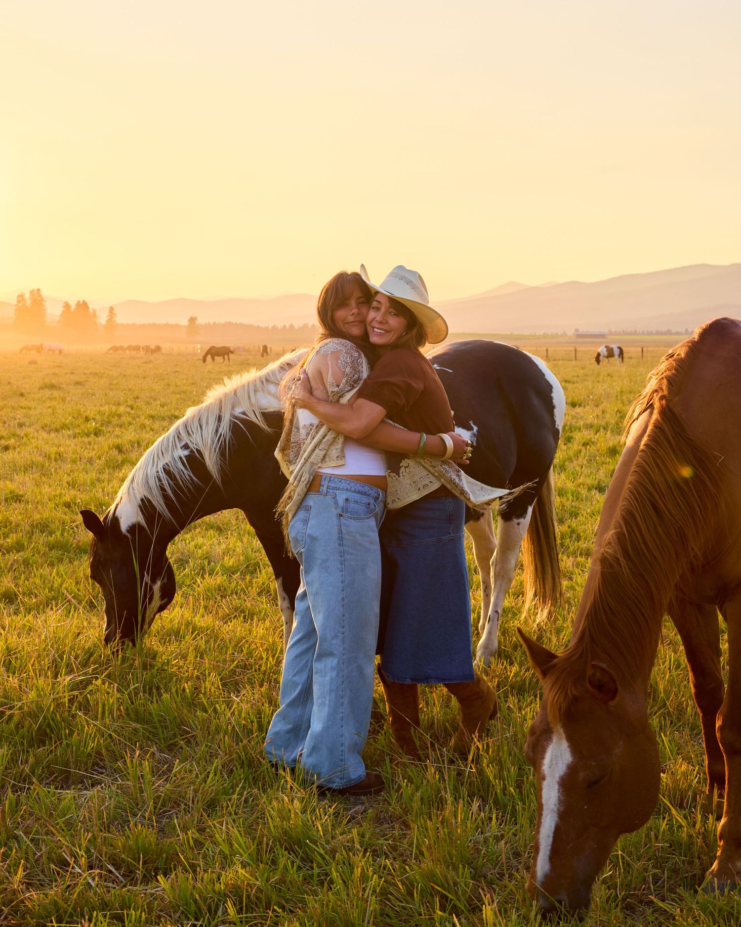 Two women hugging warmly at golden sunset in a field with horses grazing and mountains in background.