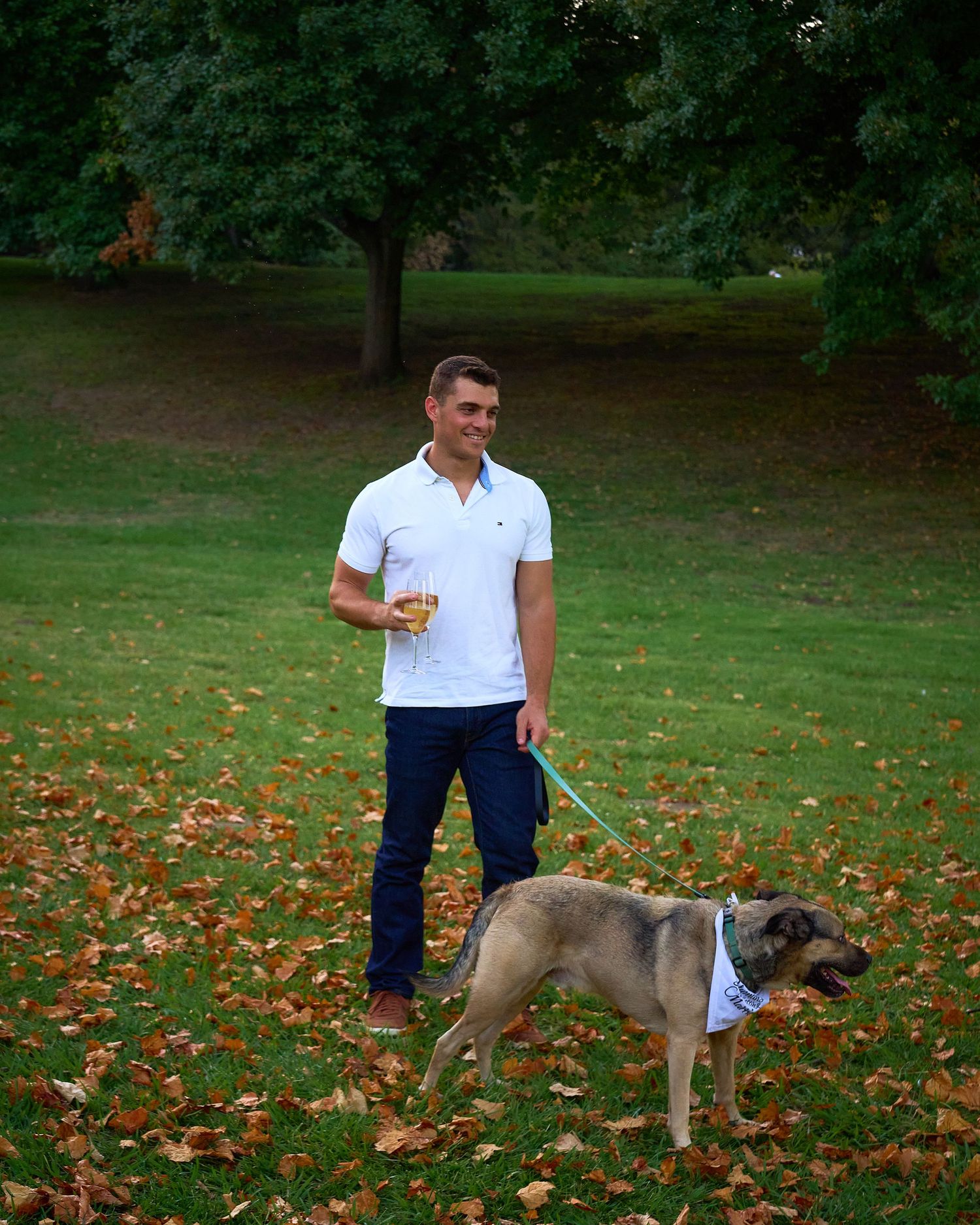 Man in white polo holding champagne glass while walking dog on leash in autumn park.