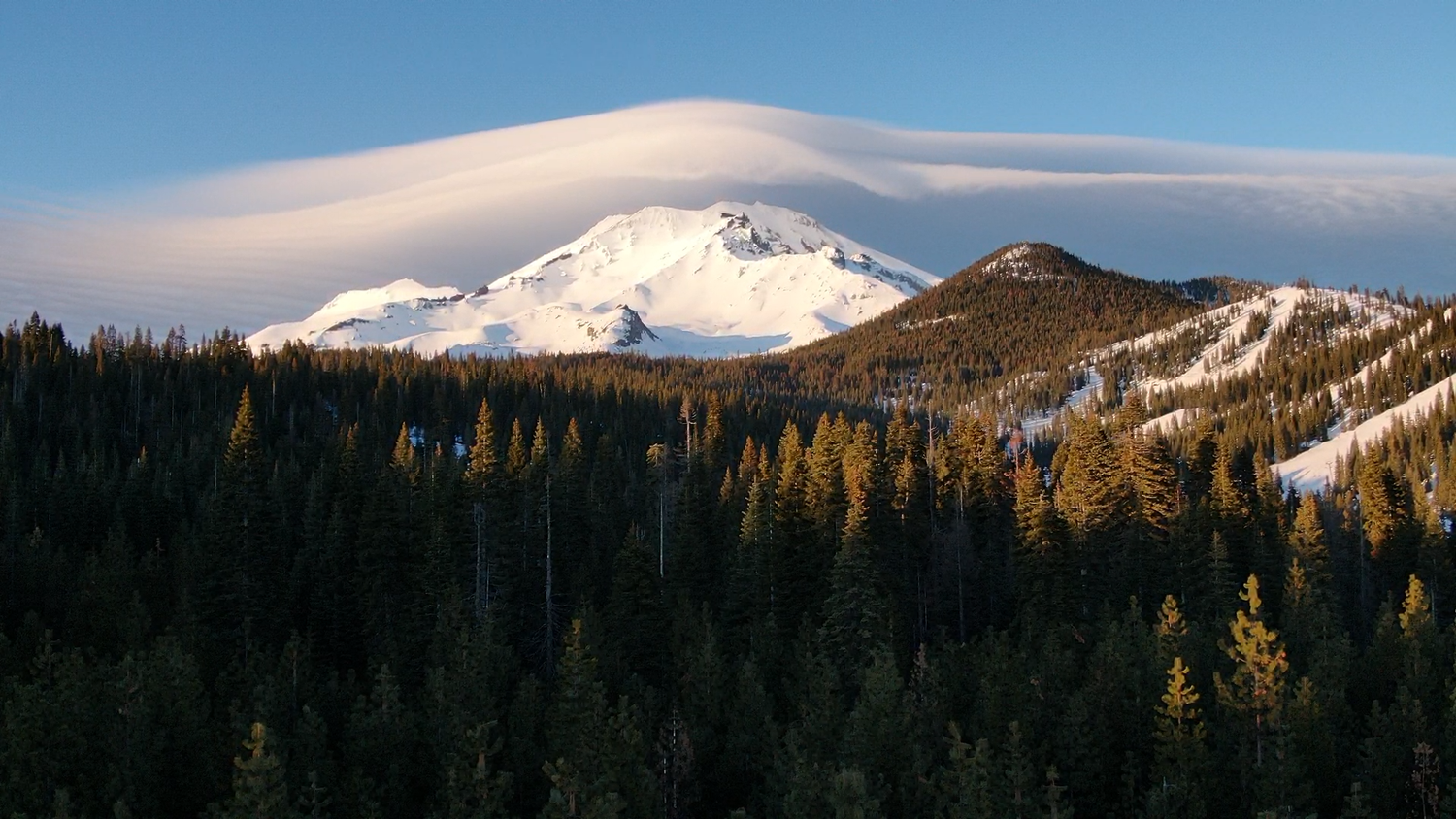Winter mountain landscape featuring a snow-capped peak with lenticular cloud above golden-lit evergreen trees.
