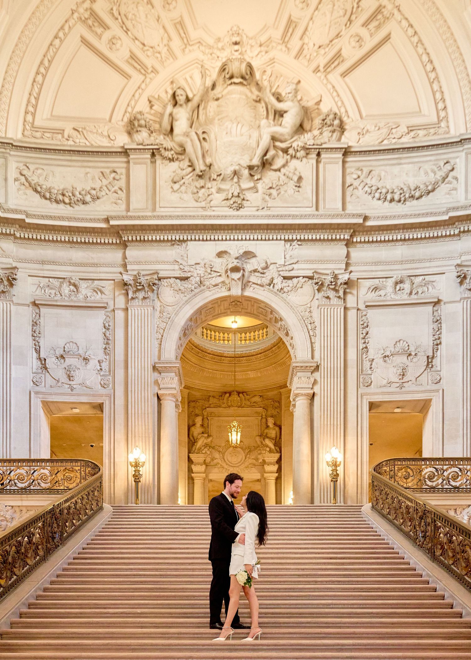 Couple sharing romantic kiss on grand staircase inside ornate San Francisco City Hall rotunda.