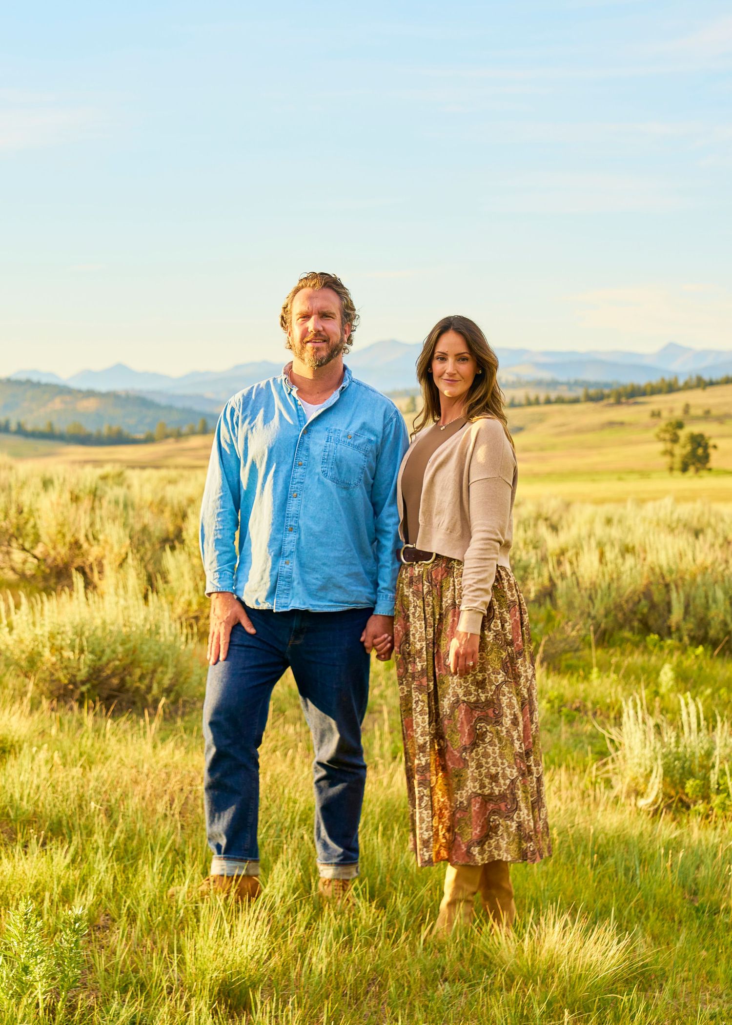 Couple holds hands smiling in sagebrush meadow with sweeping mountain valley panorama behind them.
