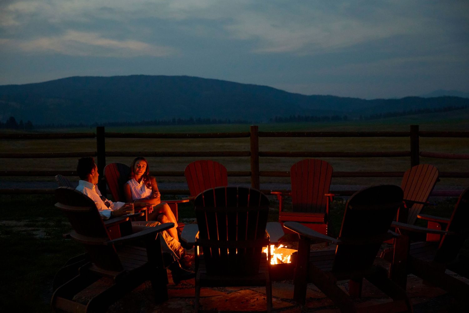 People gathered around a glowing campfire at dusk with mountain views and red Adirondack chairs.