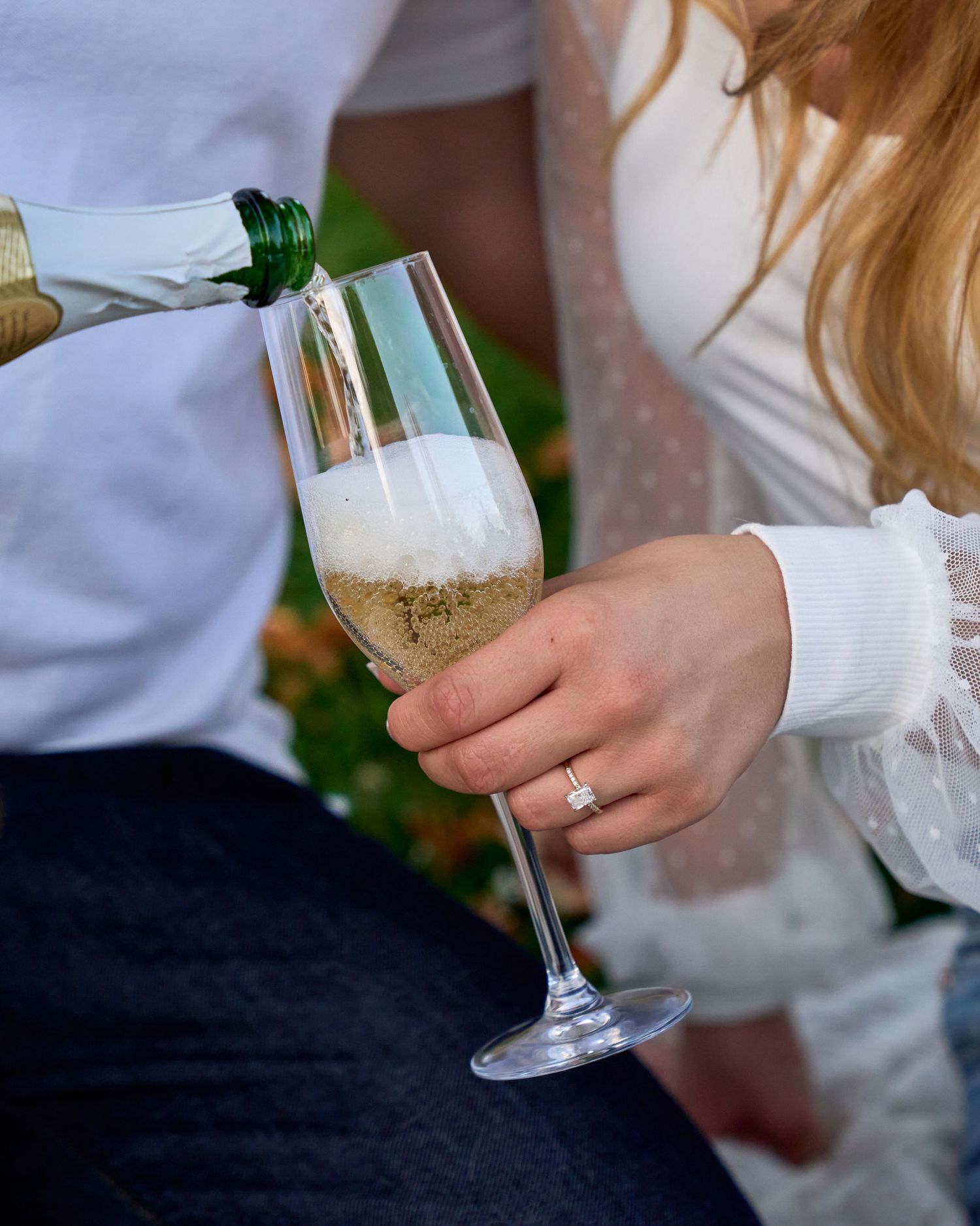 Man pouring champagne into a glass held by woman wearing an engagement ring outdoors.