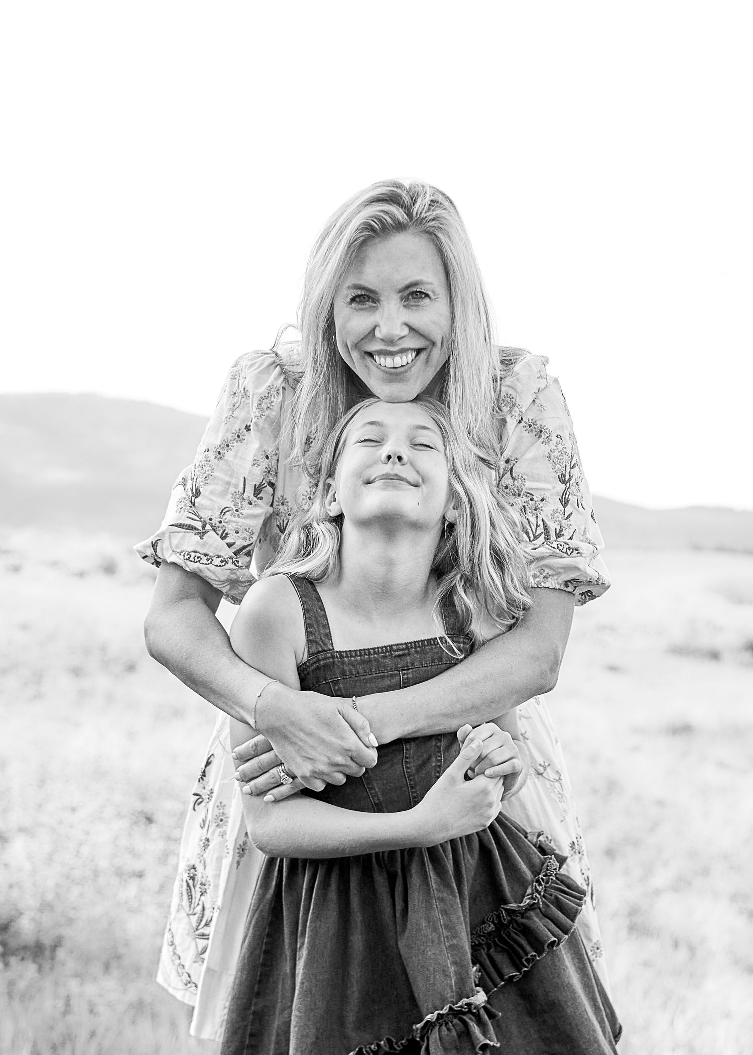 Black and white portrait of smiling woman hugging young girl from behind in an open field.