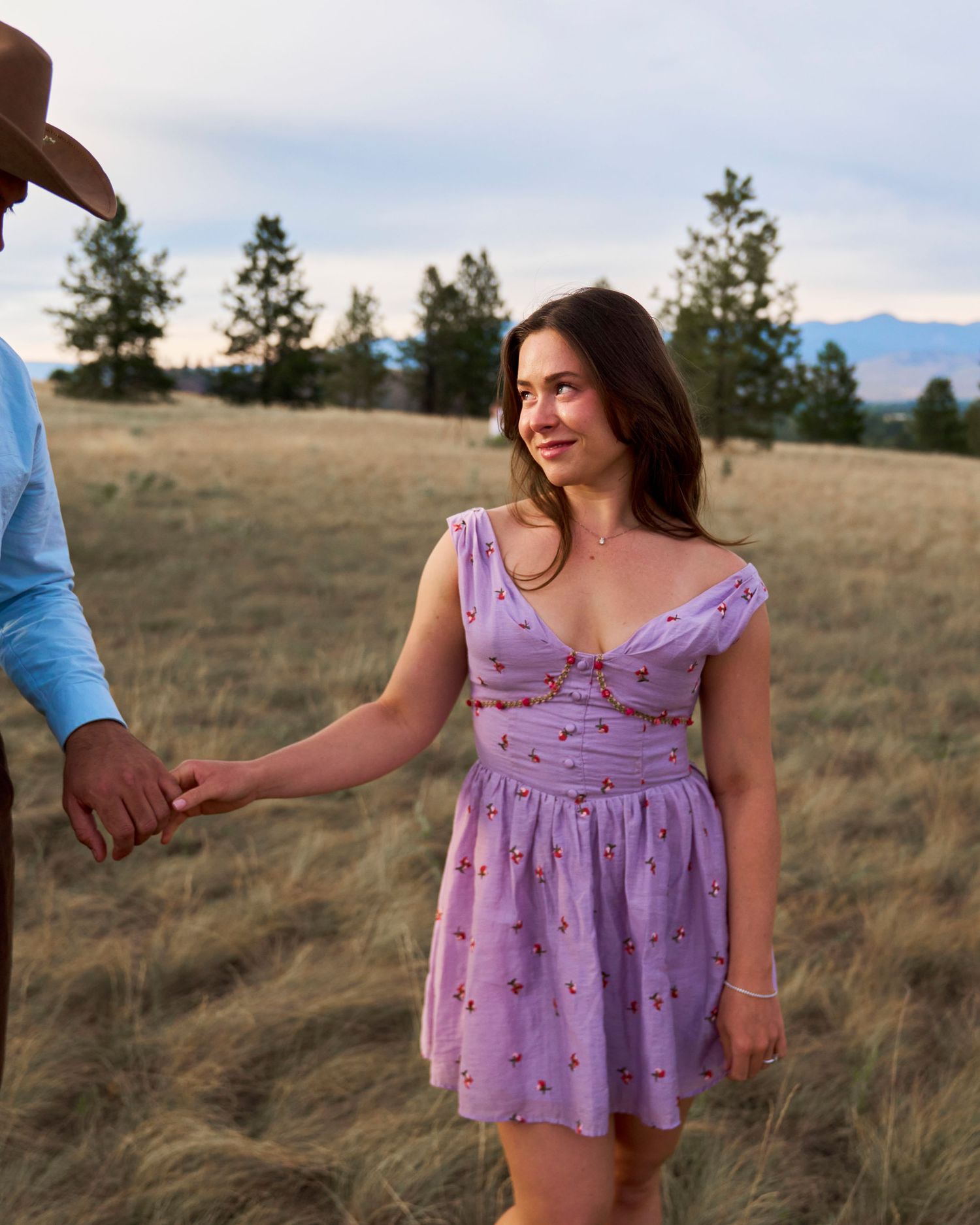 Woman in lavender floral dress holds hands with partner while walking through a golden grass field at sunset.
