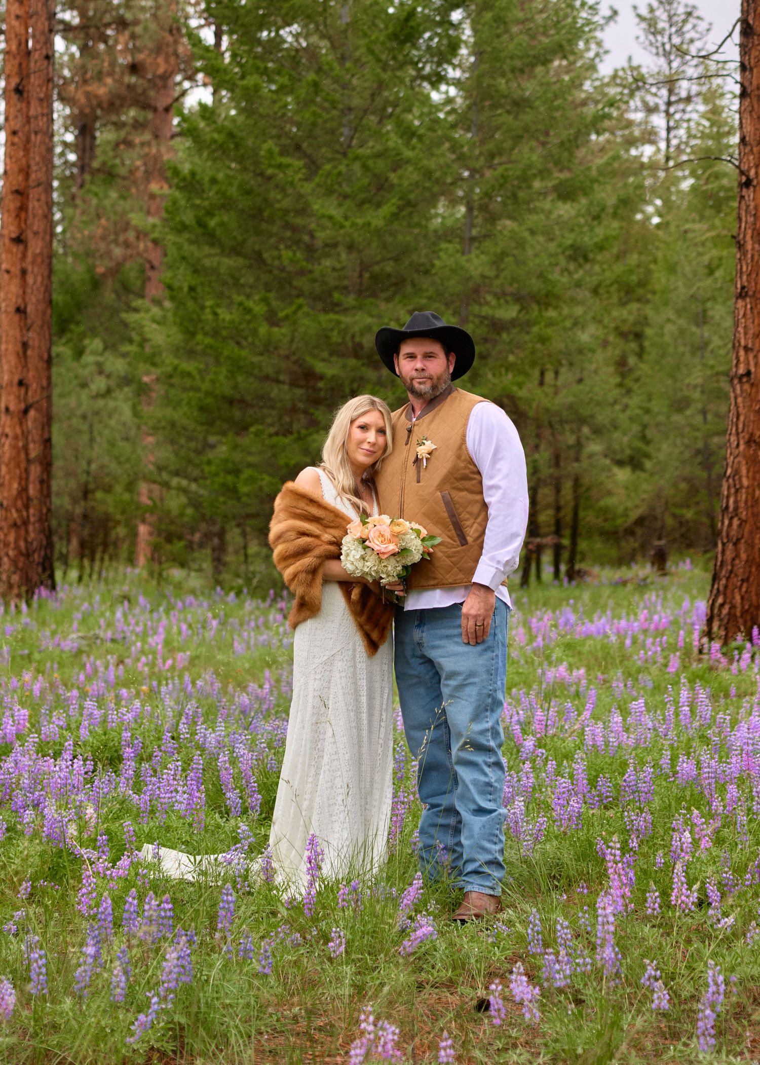Couple poses in a vibrant purple lupine meadow, groom in cowboy hat and vest, bride in lace gown with fur wrap.