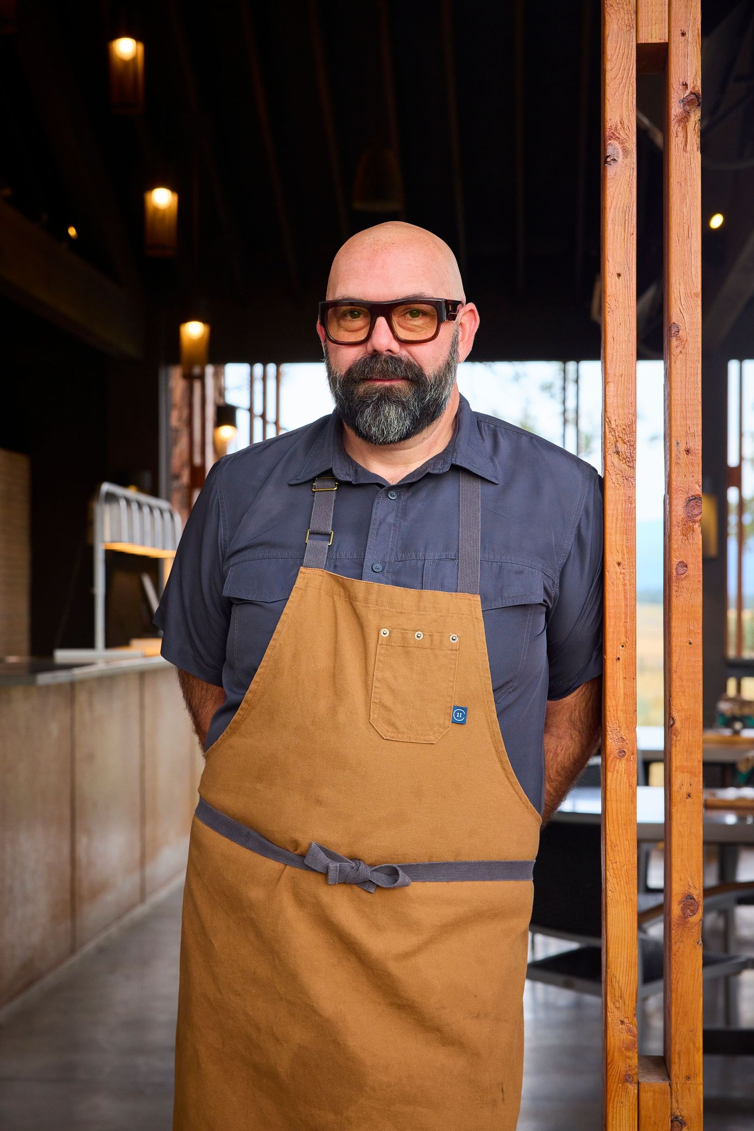 Bald bearded chef in brown apron and glasses standing in rustic restaurant interior.