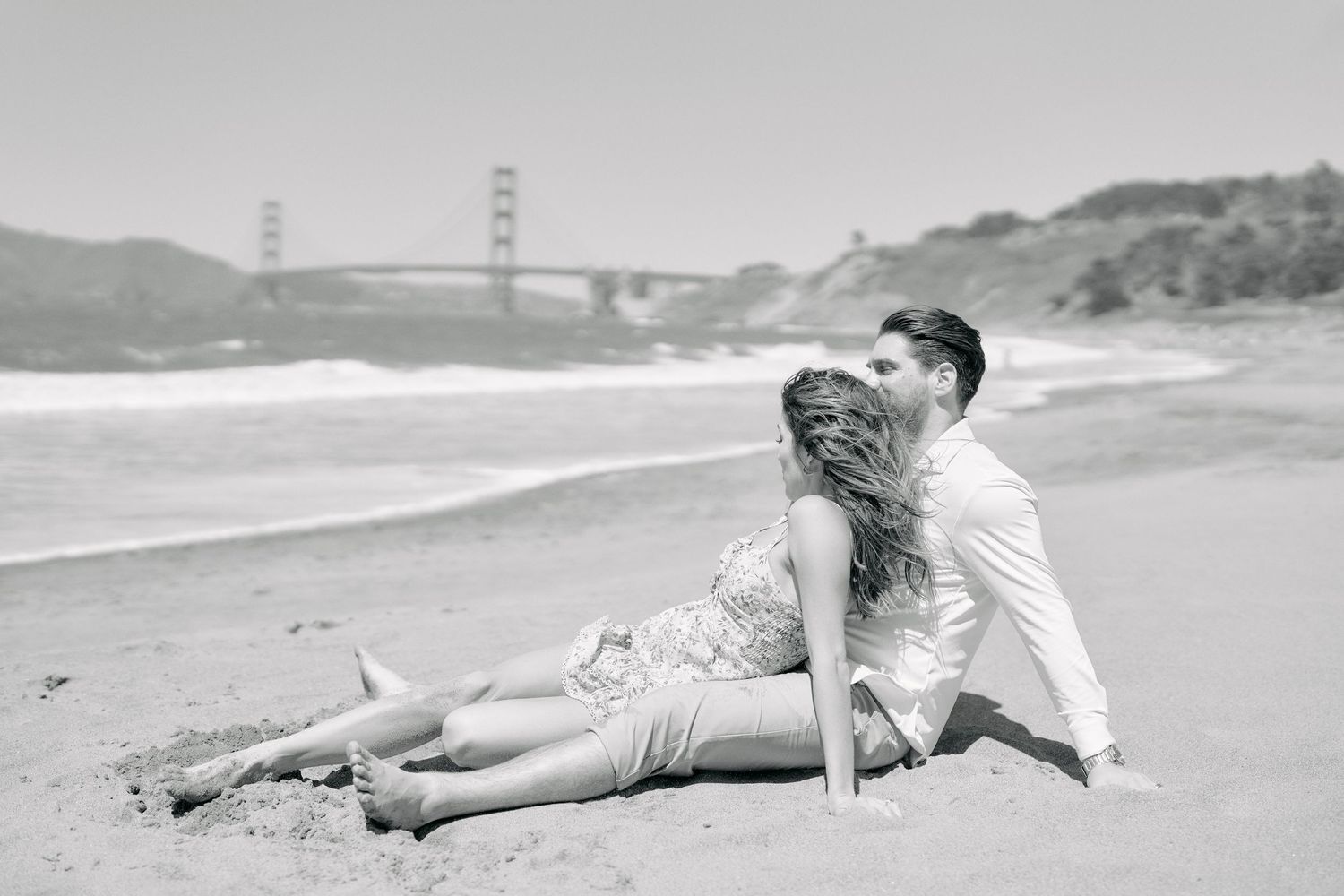 Black and white photo of a couple sitting and kissing on sandy beach with Golden Gate Bridge visible.