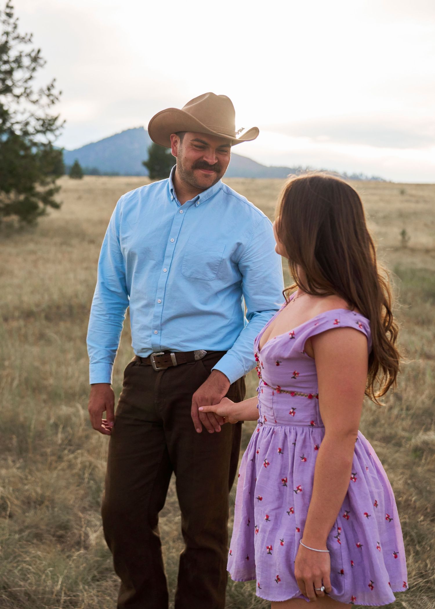 Couple holding hands and smiling at each other in a golden meadow with mountains visible in the background.