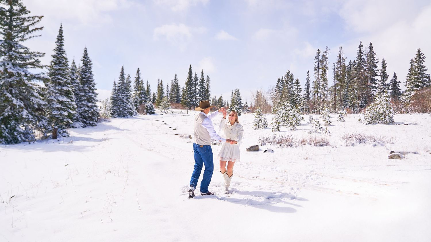 Newlywed couple dance together in open snowy field surrounded by snow-covered evergreen trees.