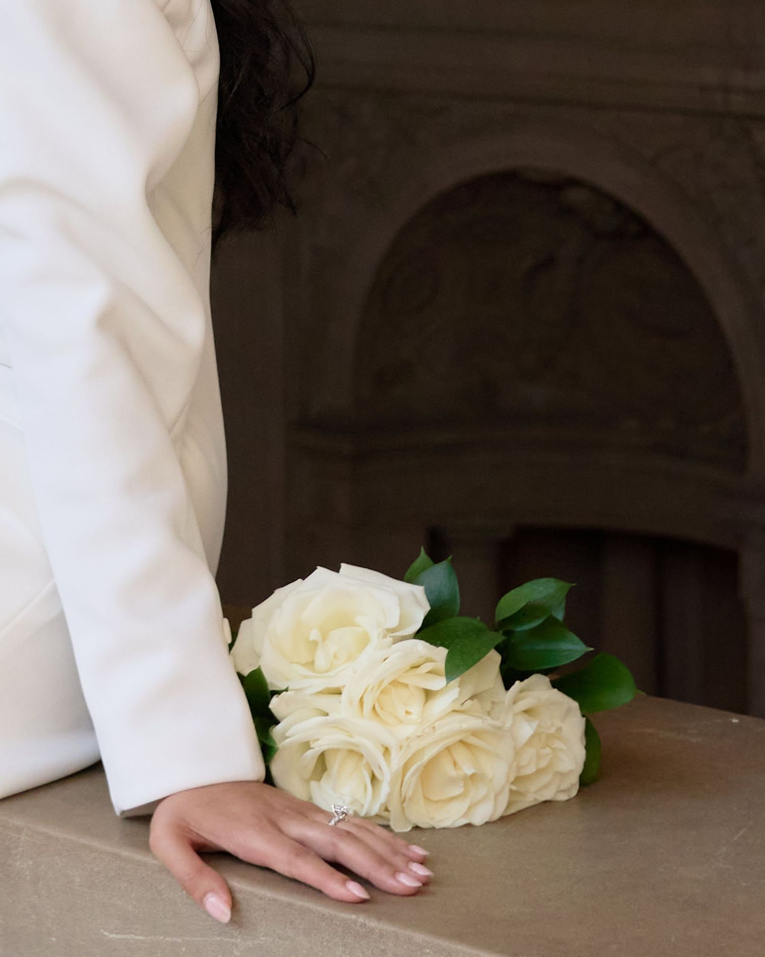 Bride holds elegant bouquet of white roses against stone ledge wearing white blazer wedding dress.