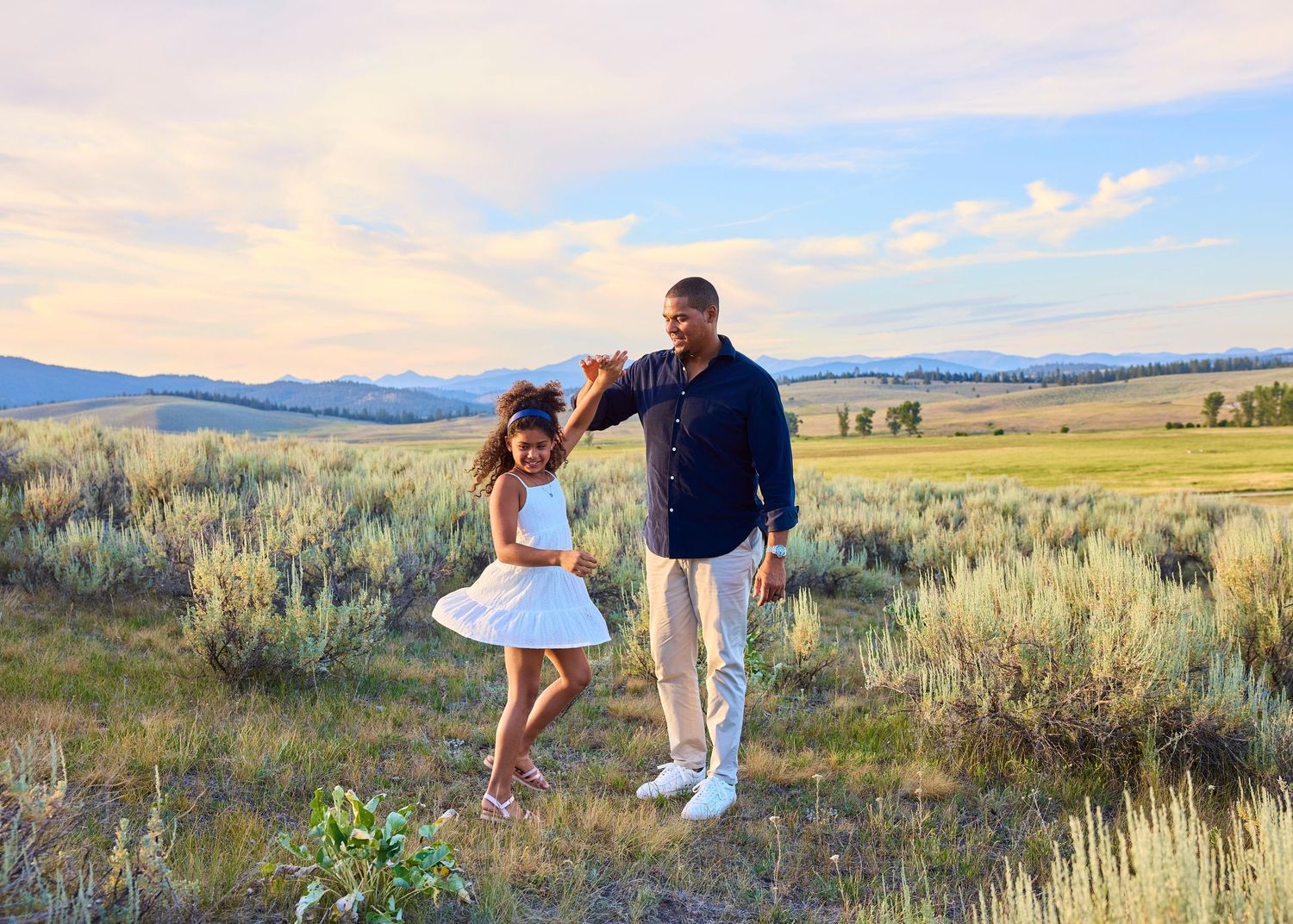 A father twirls his young daughter in a white dress across a scenic sagebrush meadow at golden hour.